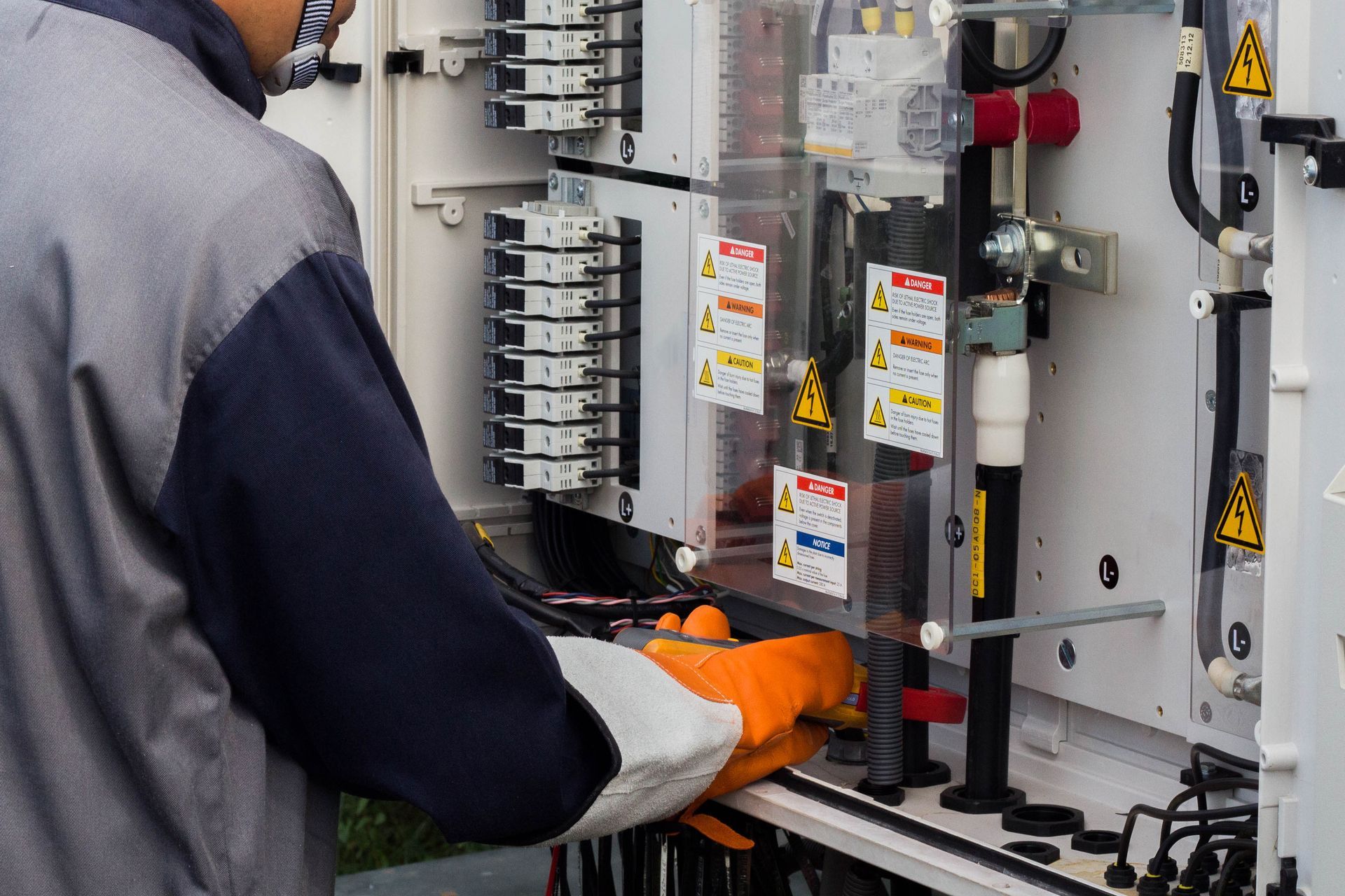 A technician wearing orange protective gloves works on the internal electrical wiring of a power distribution cabinet.