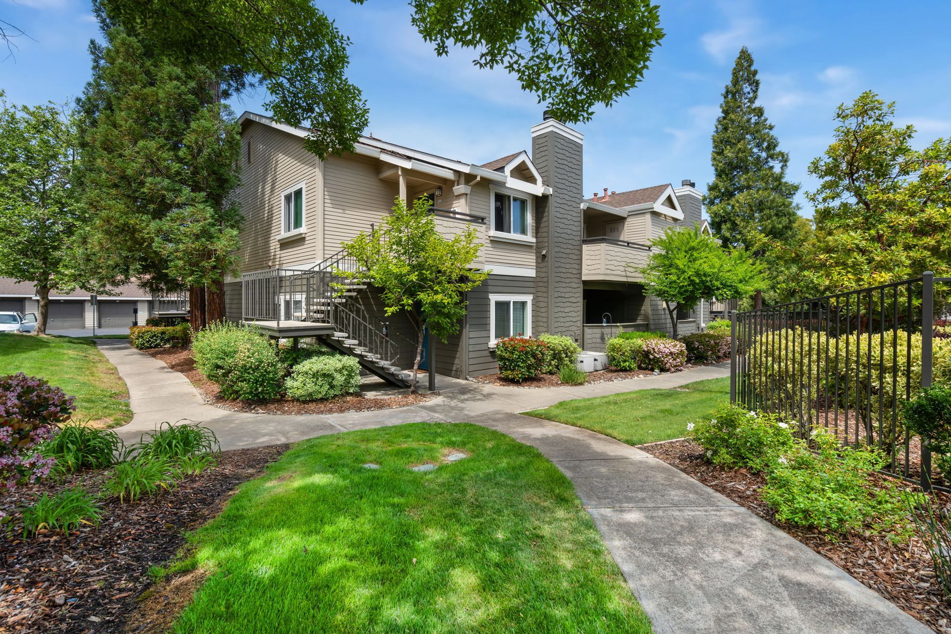 Exterior view of a beige two-story apartment building with landscaped paths and stairs surrounded by trees