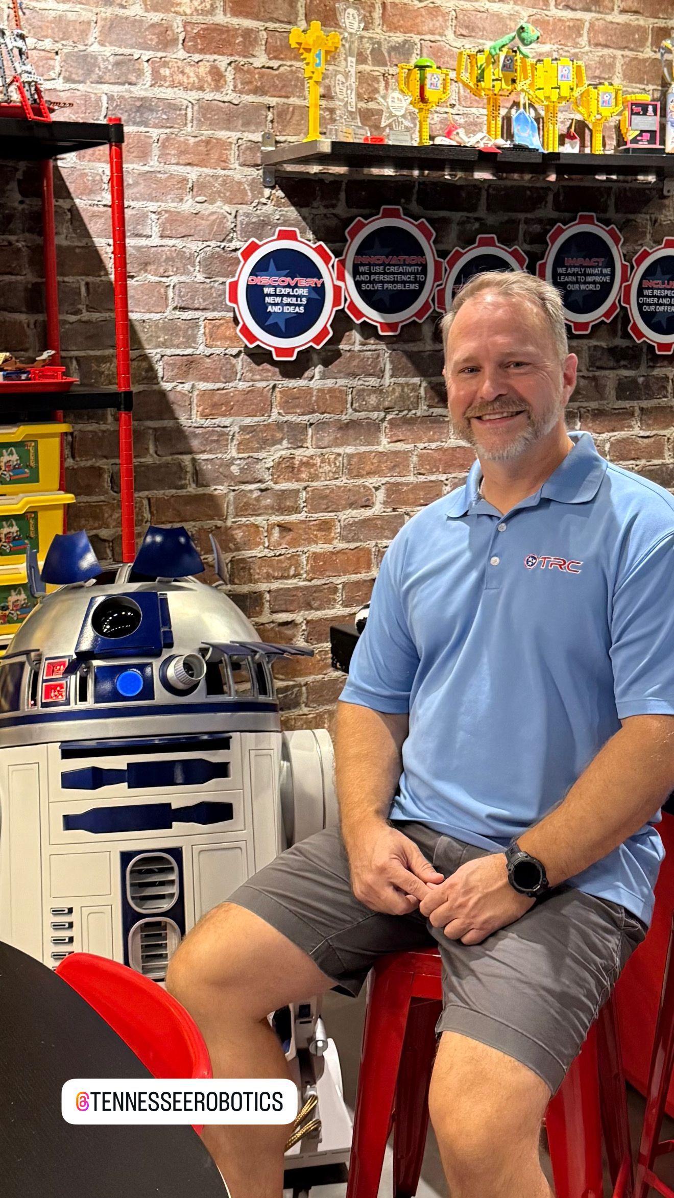 Coach Mike in blue shirt, sitting with R2-D2. Red bar stool, brick wall, and LEGO displays in background.