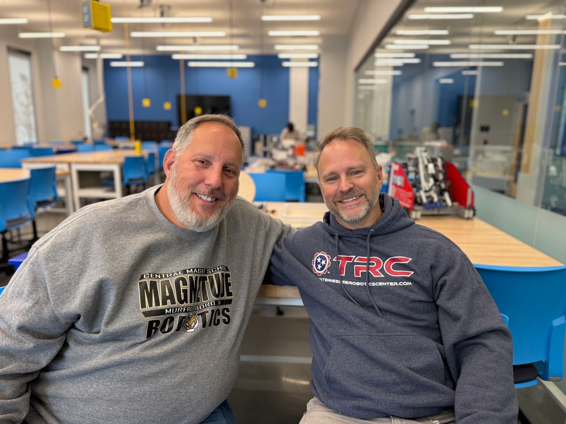 Coach Mike and Coach Mark, leaning on a table in a workshop. One wears a gray sweatshirt, the other a blue hoodie.