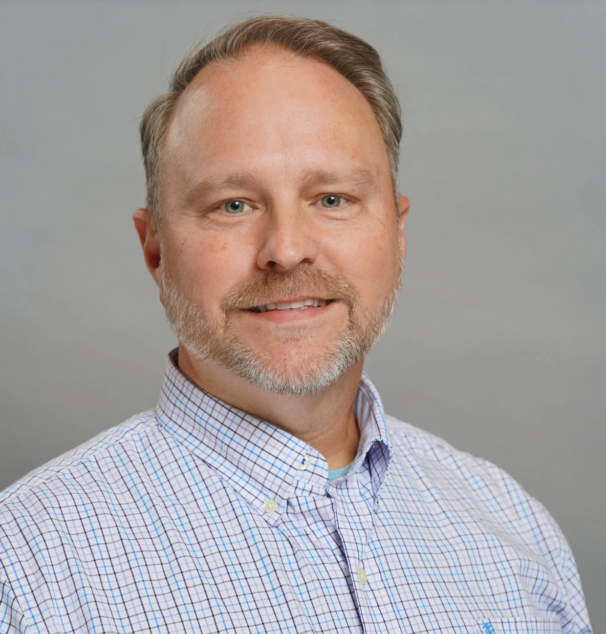 A man with a beard is wearing a plaid shirt and smiling for the camera.