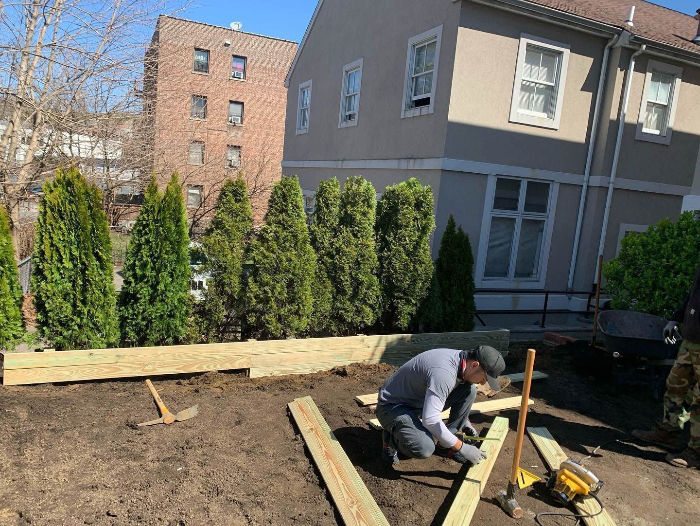 Man building a wooden structure outdoors. Trees and a building are in the background. Sunny day.