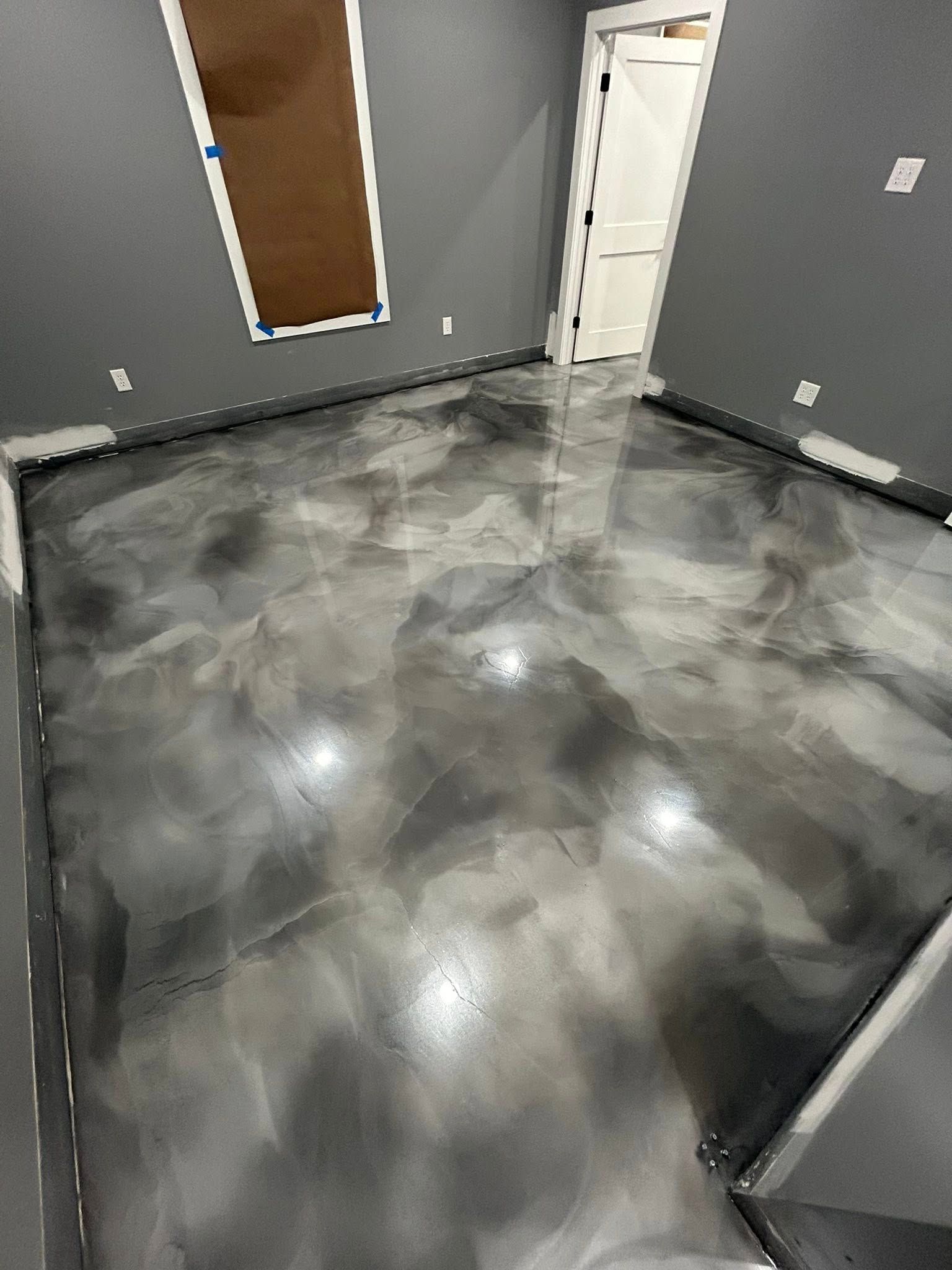 A room with a polished, grey-toned metallic epoxy floor, dark grey walls, and a white door frame.