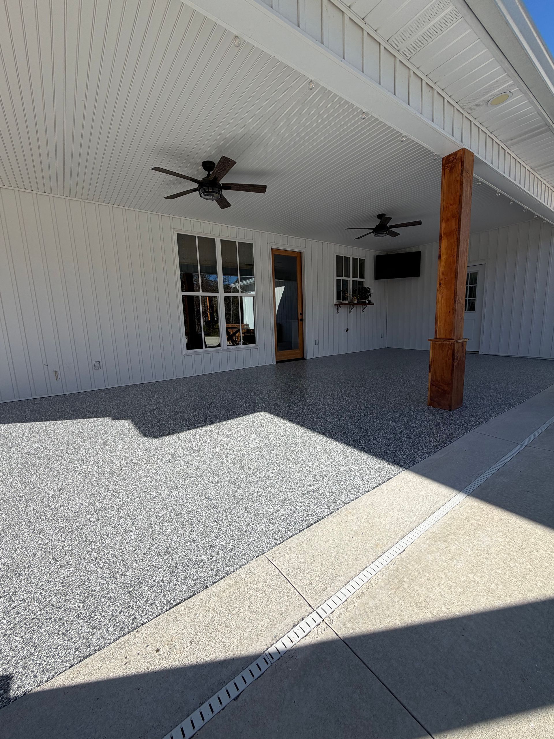 A covered porch with a gray-and-white flecked epoxy floor, white siding, two ceiling fans, and a rustic wooden support post.