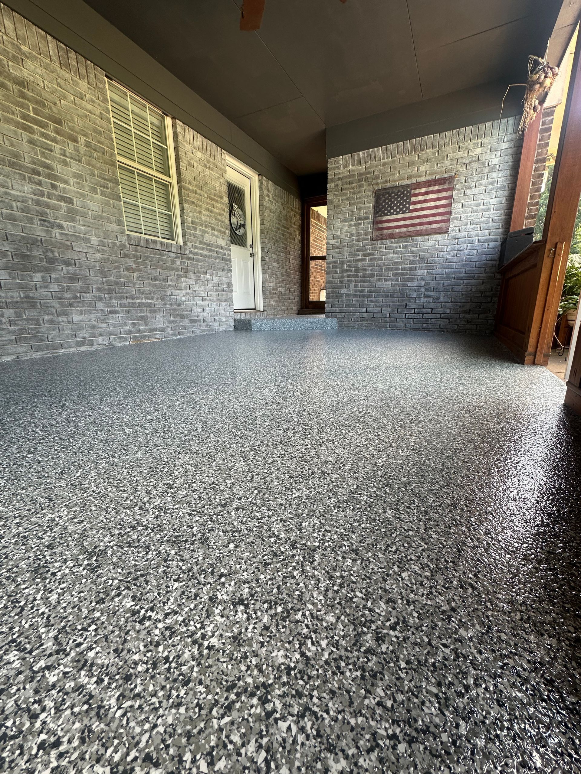 A covered porch with a high-gloss, gray speckled epoxy floor, brick walls, and an American flag wall decoration.