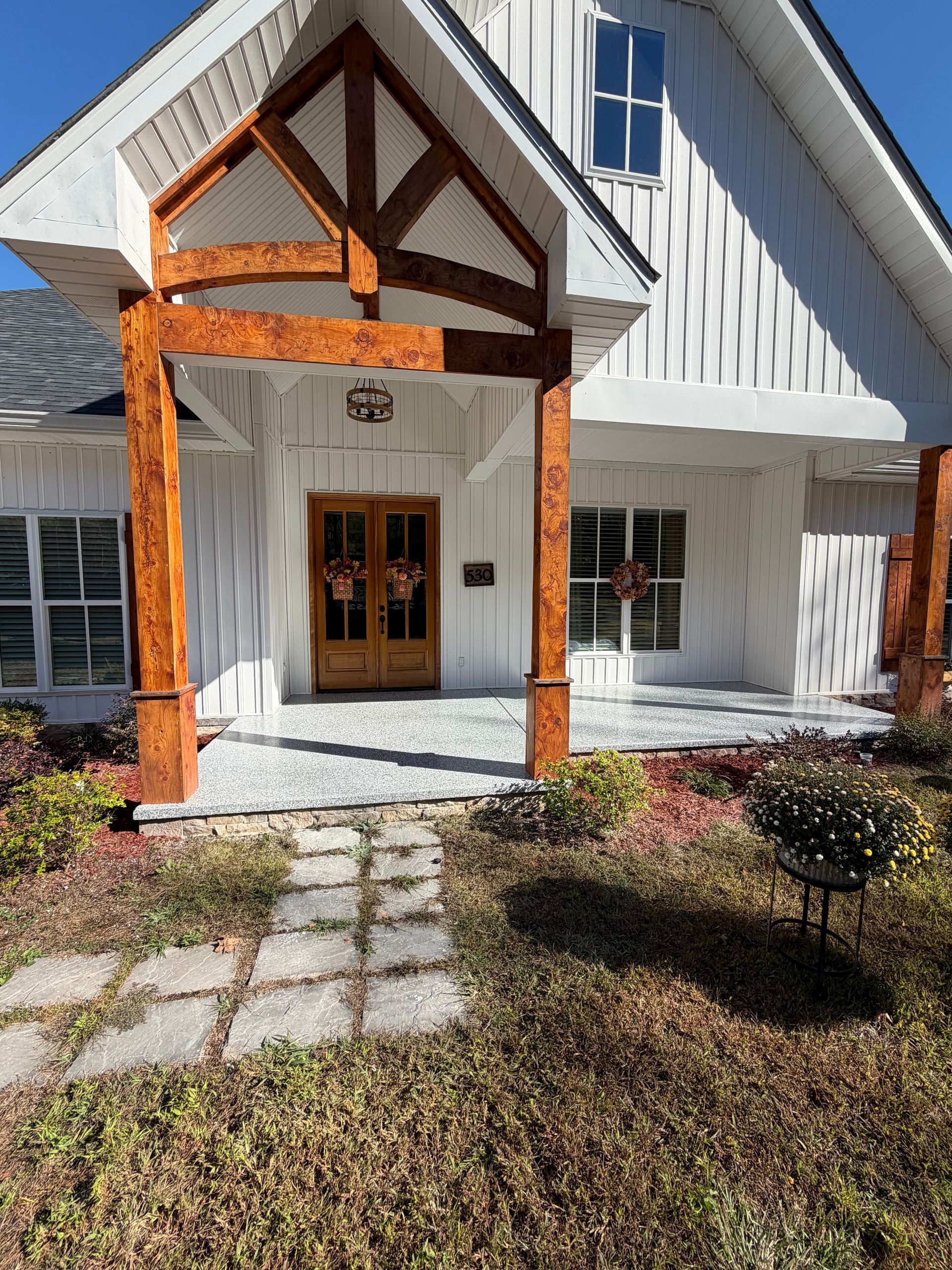 White farmhouse with wood beam porch, double doors, and stone walkway under a clear blue sky.