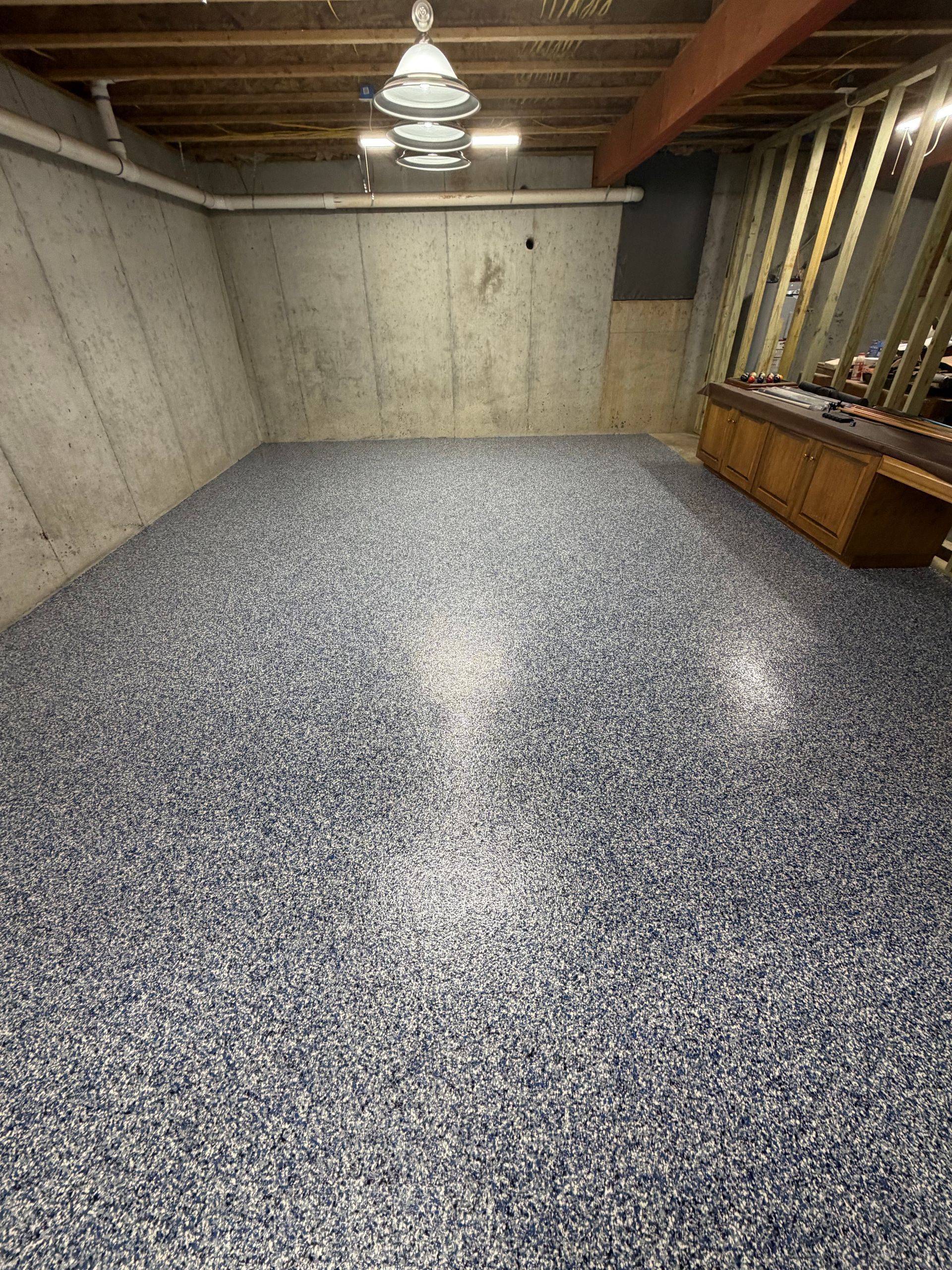 A basement with a freshly applied gray and blue speckled epoxy floor, concrete walls, and unfinished wooden wall framing.