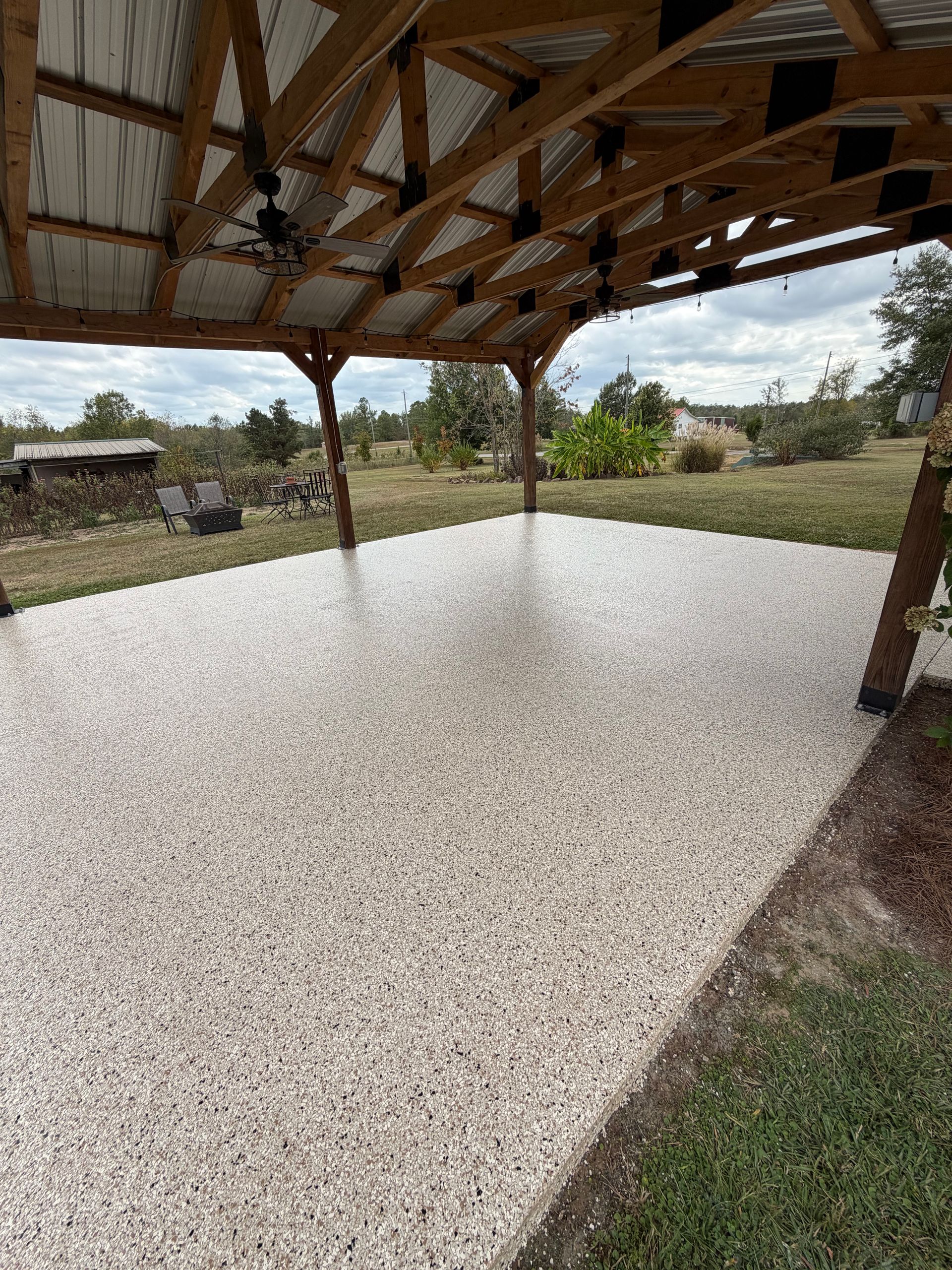 A light-colored, speckled patio surface under a wooden open-air pavilion structure in a grassy outdoor setting.