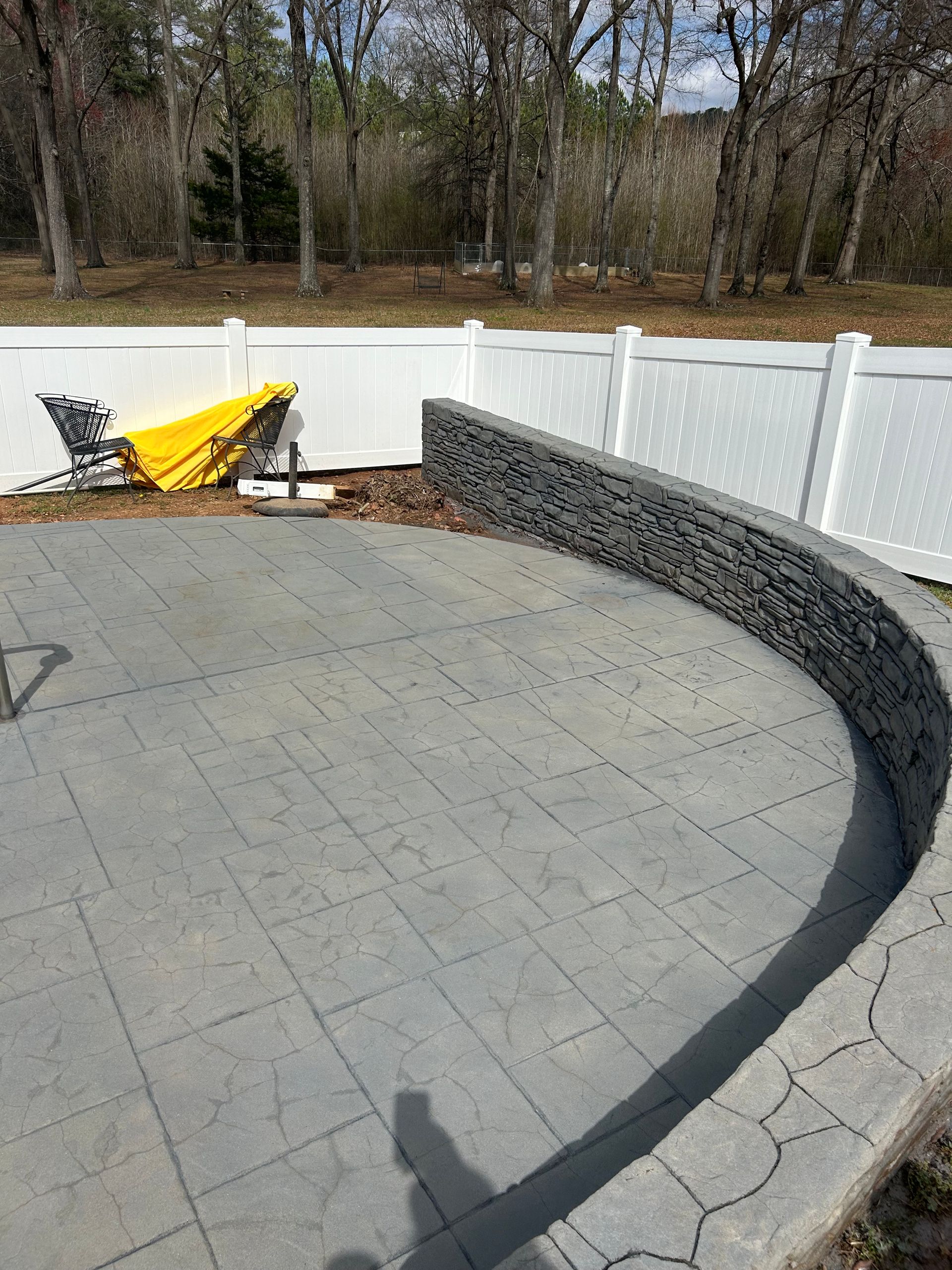 A curved stone wall sits on a gray patterned patio near a white fence, with a folded yellow umbrella in the background.