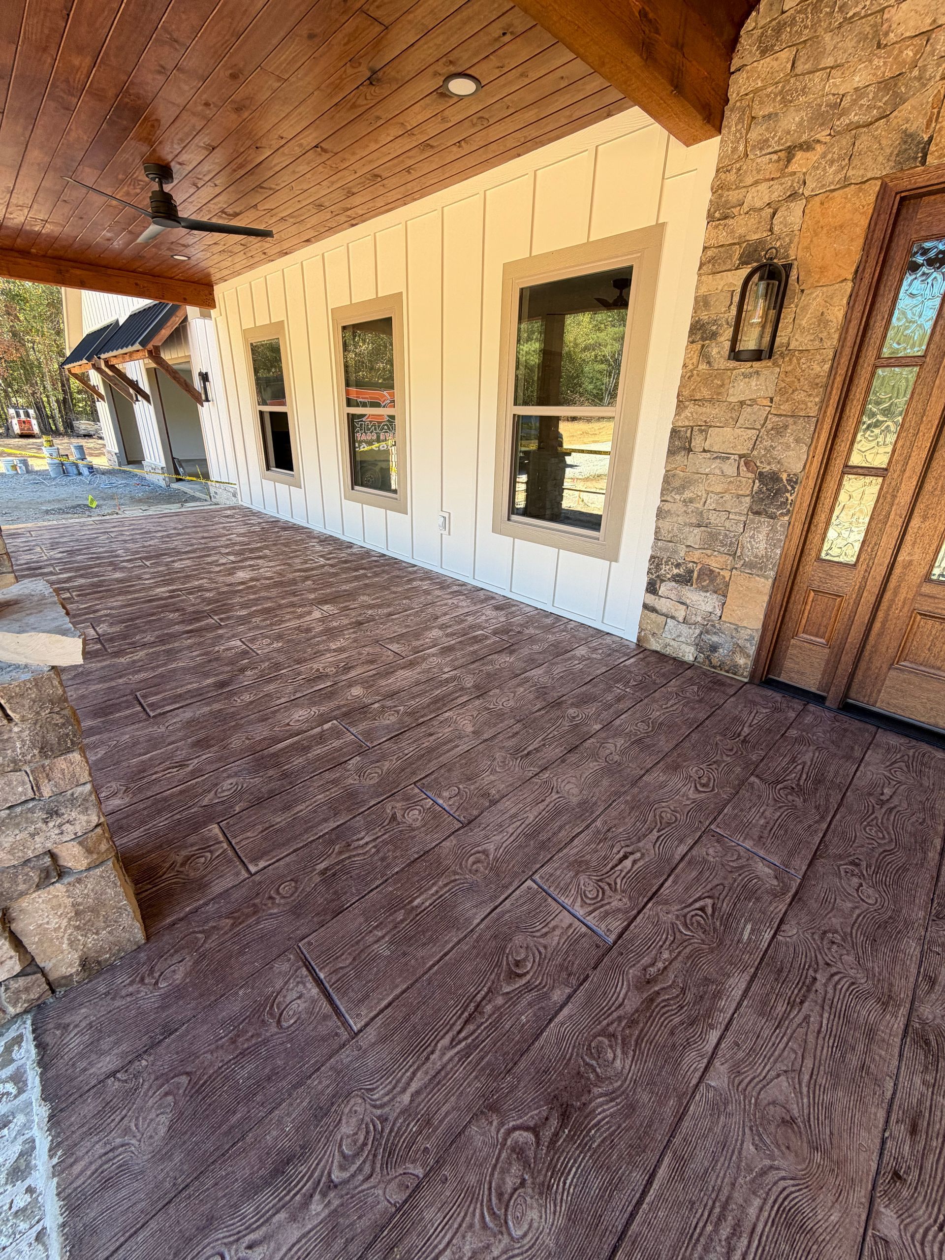 A porch with wood-look stamped concrete flooring, light-colored board and batten siding, stone accents, and a wood door.