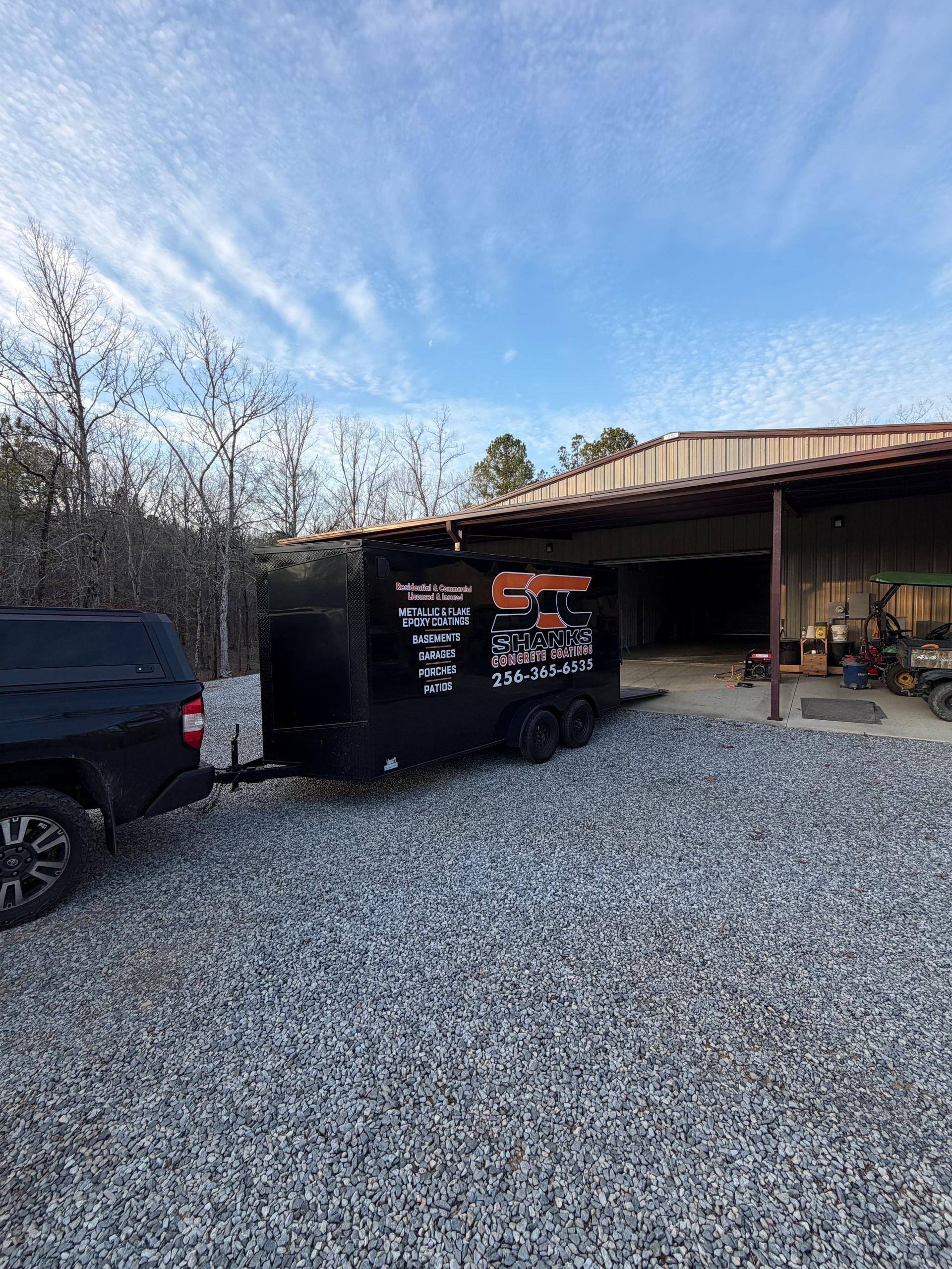 A black trailer with company branding is hitched to a pickup truck, parked on a gravel lot under a metal shed structure.