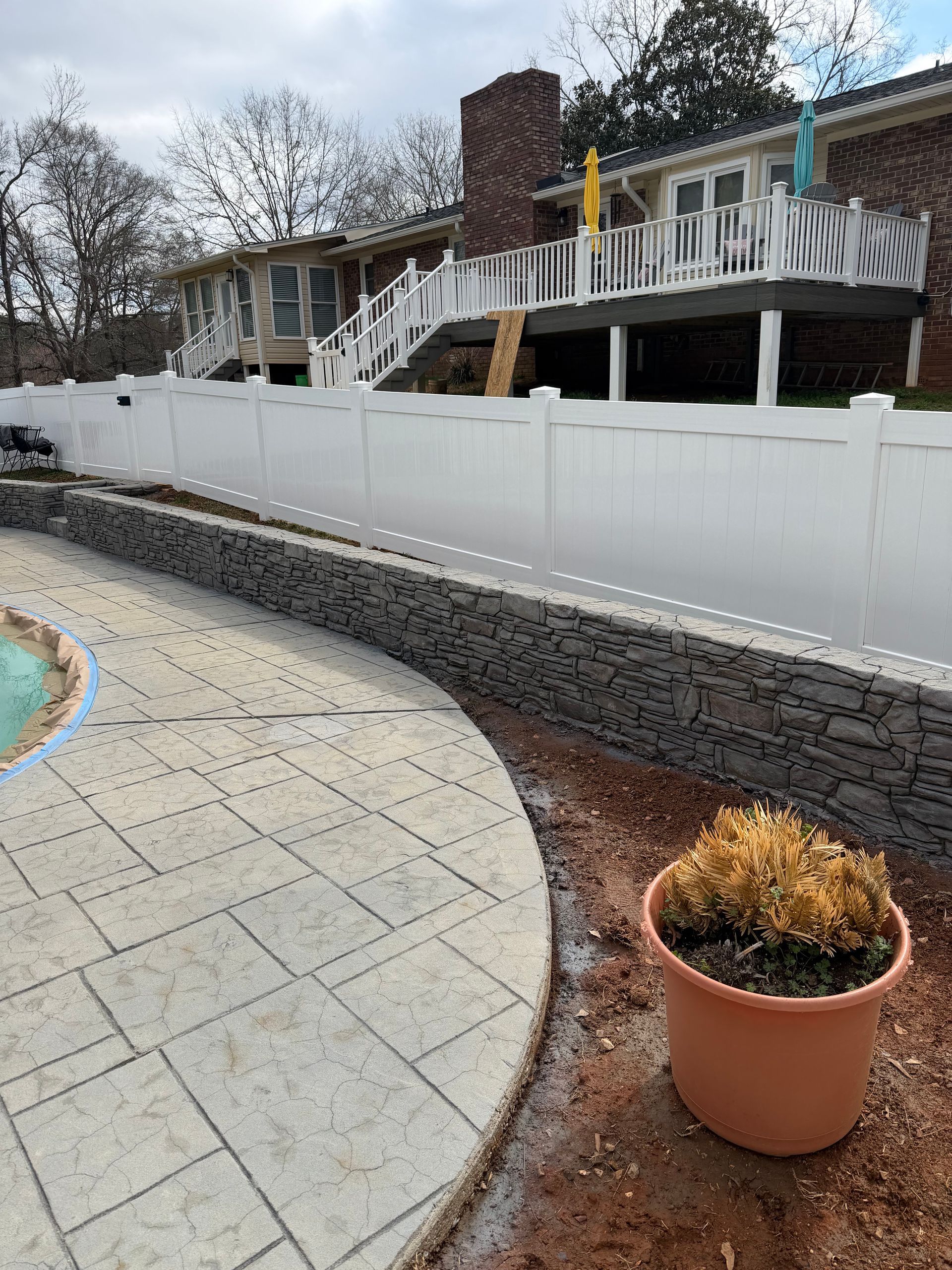 A backyard swimming pool area with a stone retaining wall, a white vinyl fence, and a wooden deck on a brick house.
