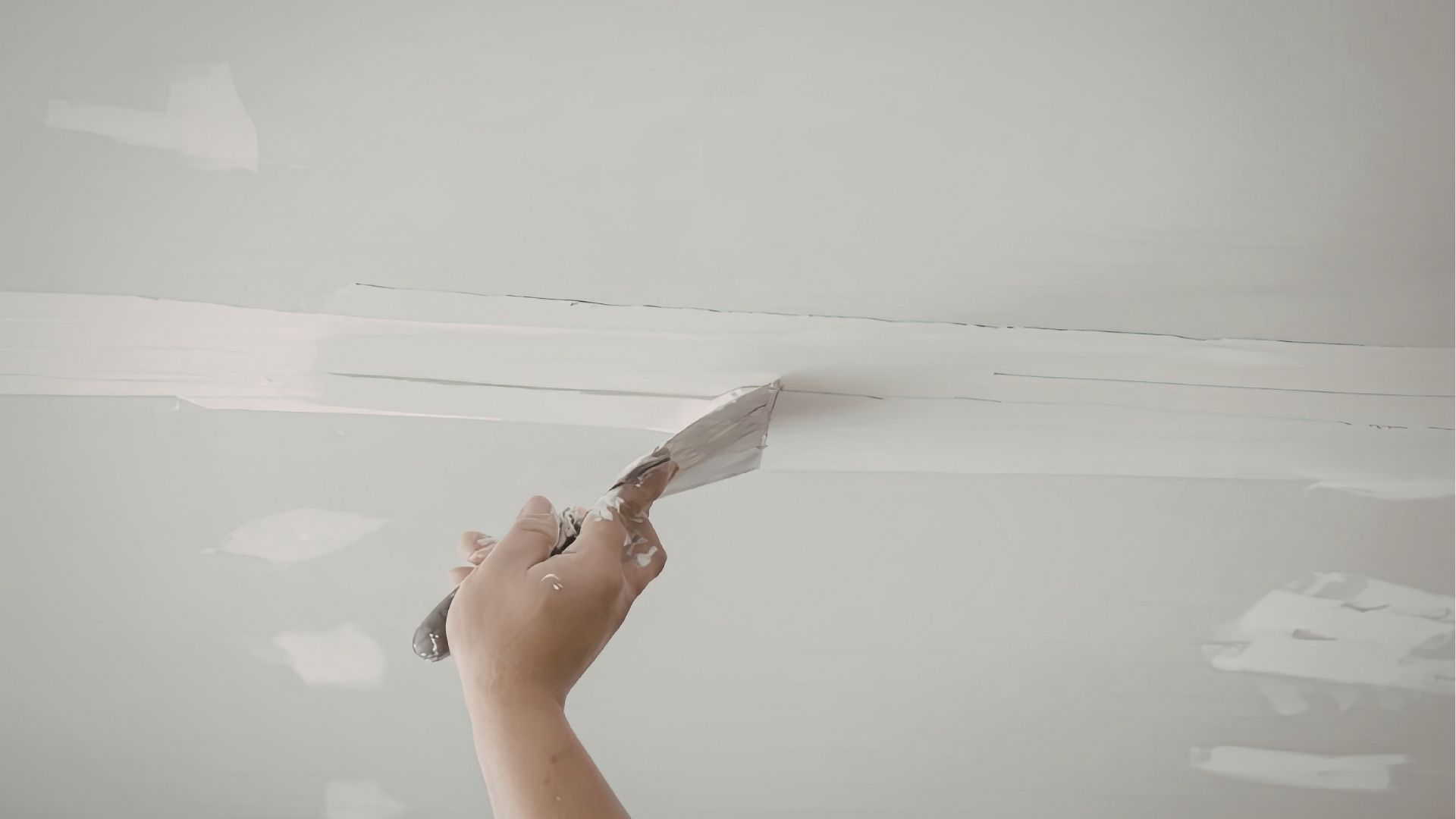 A hand uses a metal putty knife to apply joint compound to a seam on a white ceiling.