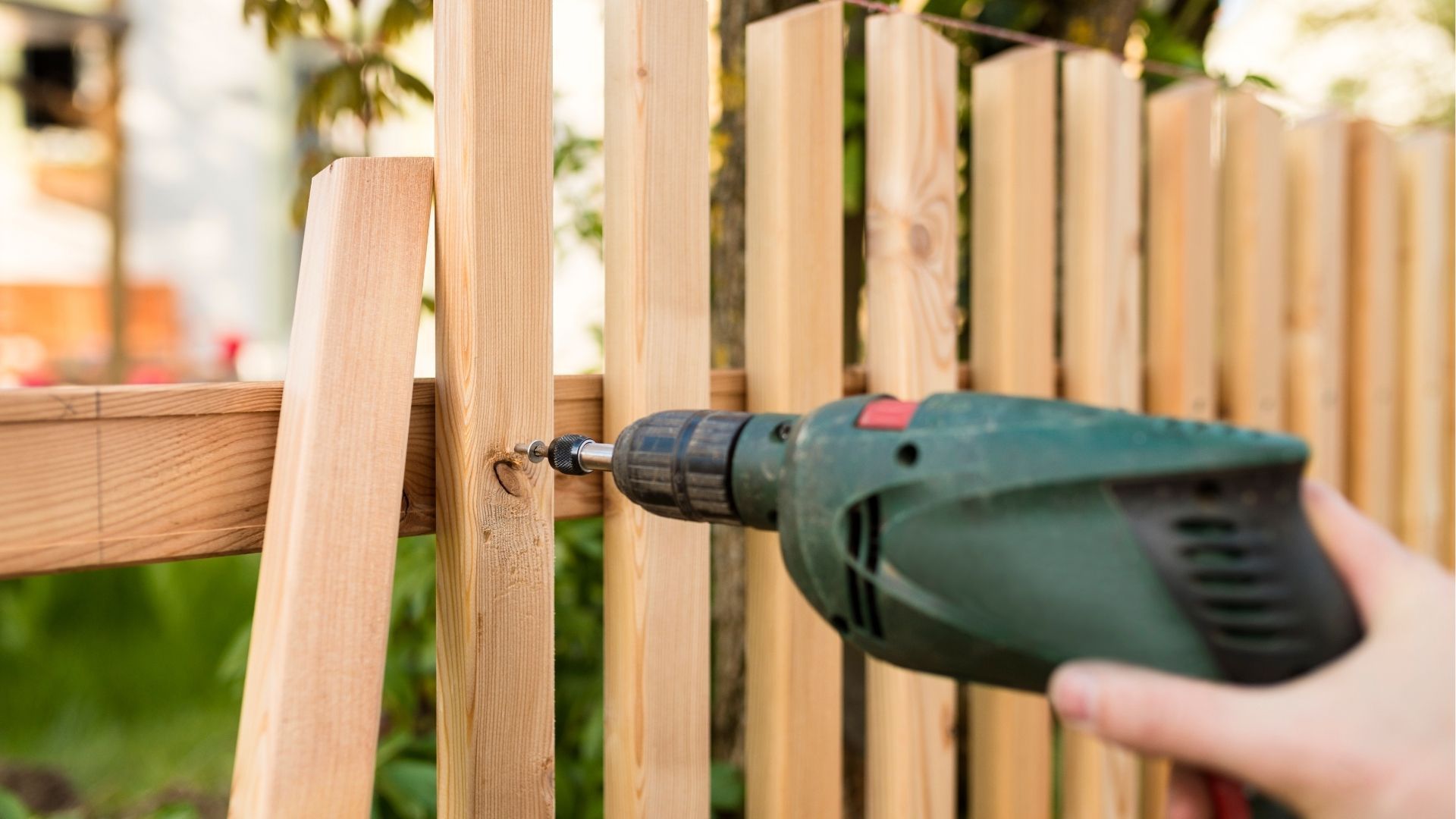 A hand uses a green power drill to attach a vertical wooden slat to a horizontal rail on a fence.