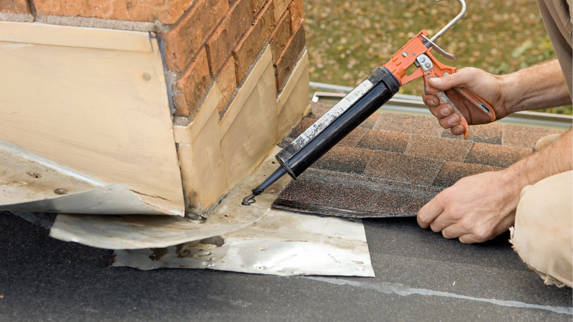 A person uses a caulk gun to apply sealant at the base of a brick chimney on a roof near overlapping shingles.