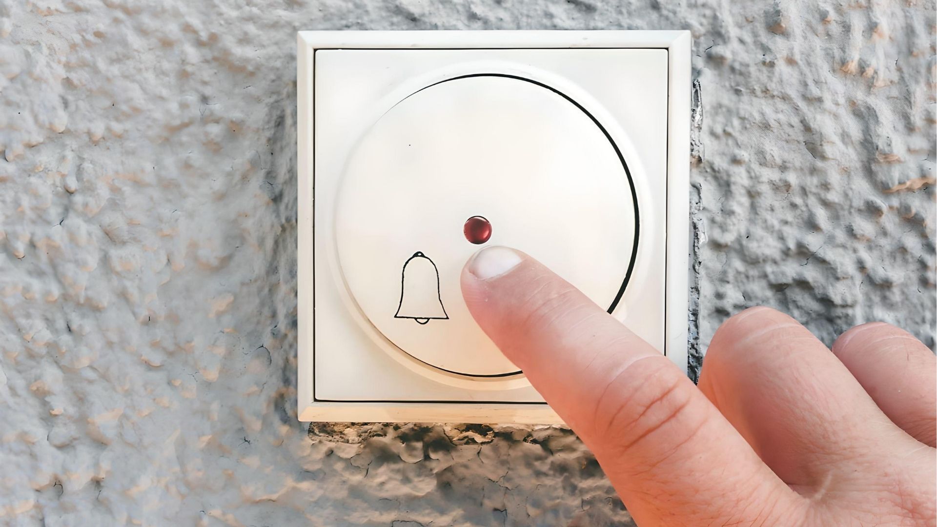 A finger presses a round, white doorbell button with a bell icon and a small red light on a textured grey wall.