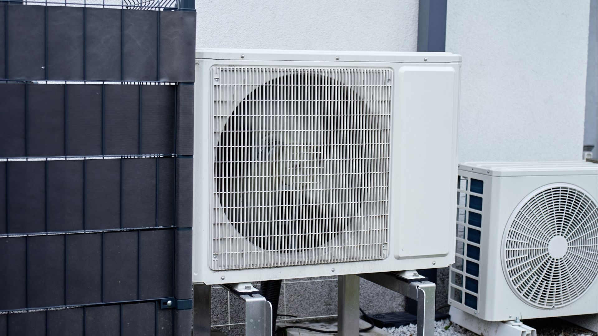 Two white heat pump outdoor units placed side-by-side on metal stands next to a tall, dark gray privacy fence.