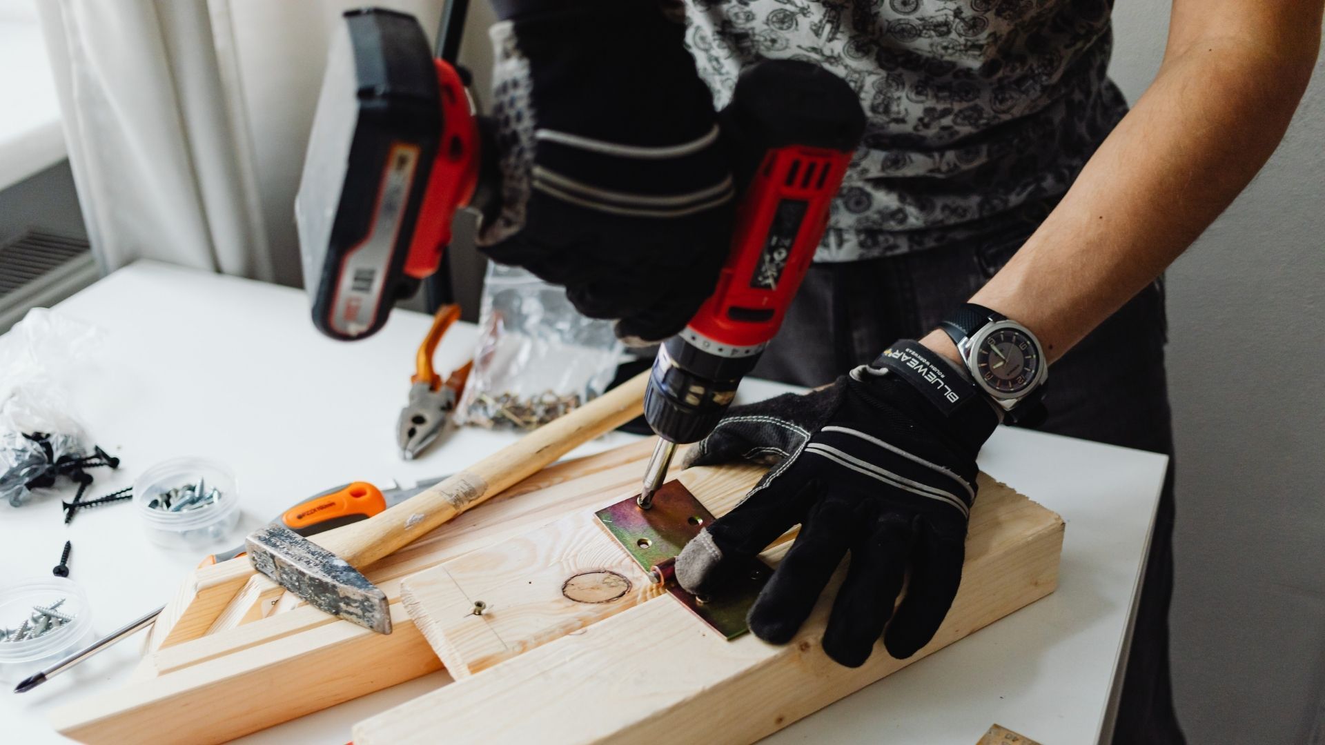 A person wearing black work gloves uses a red cordless drill to attach a metal hinge to a wooden board on a white table.