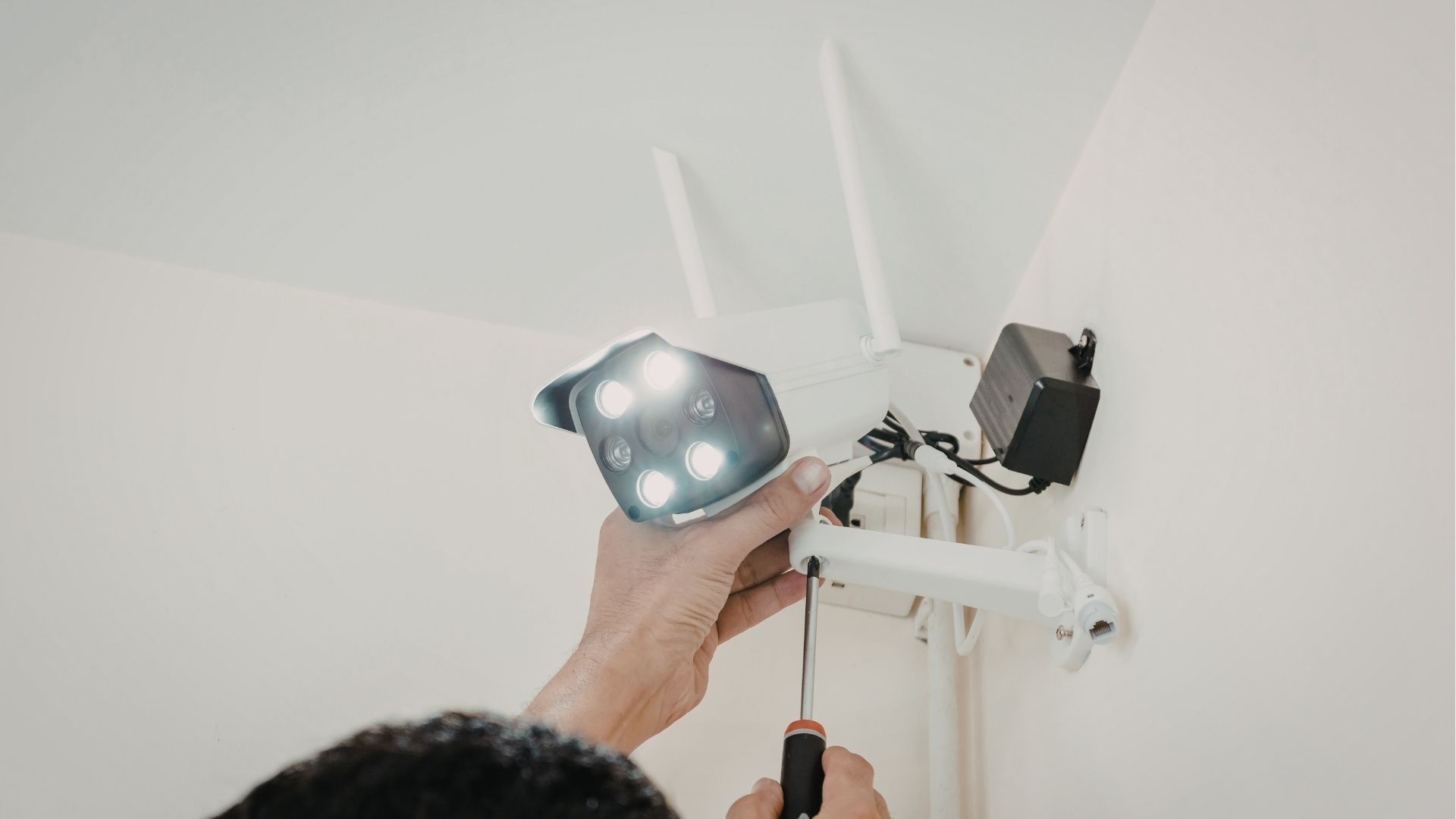 A person uses a screwdriver to mount a white, dual-antenna security camera onto a corner of a wall.
