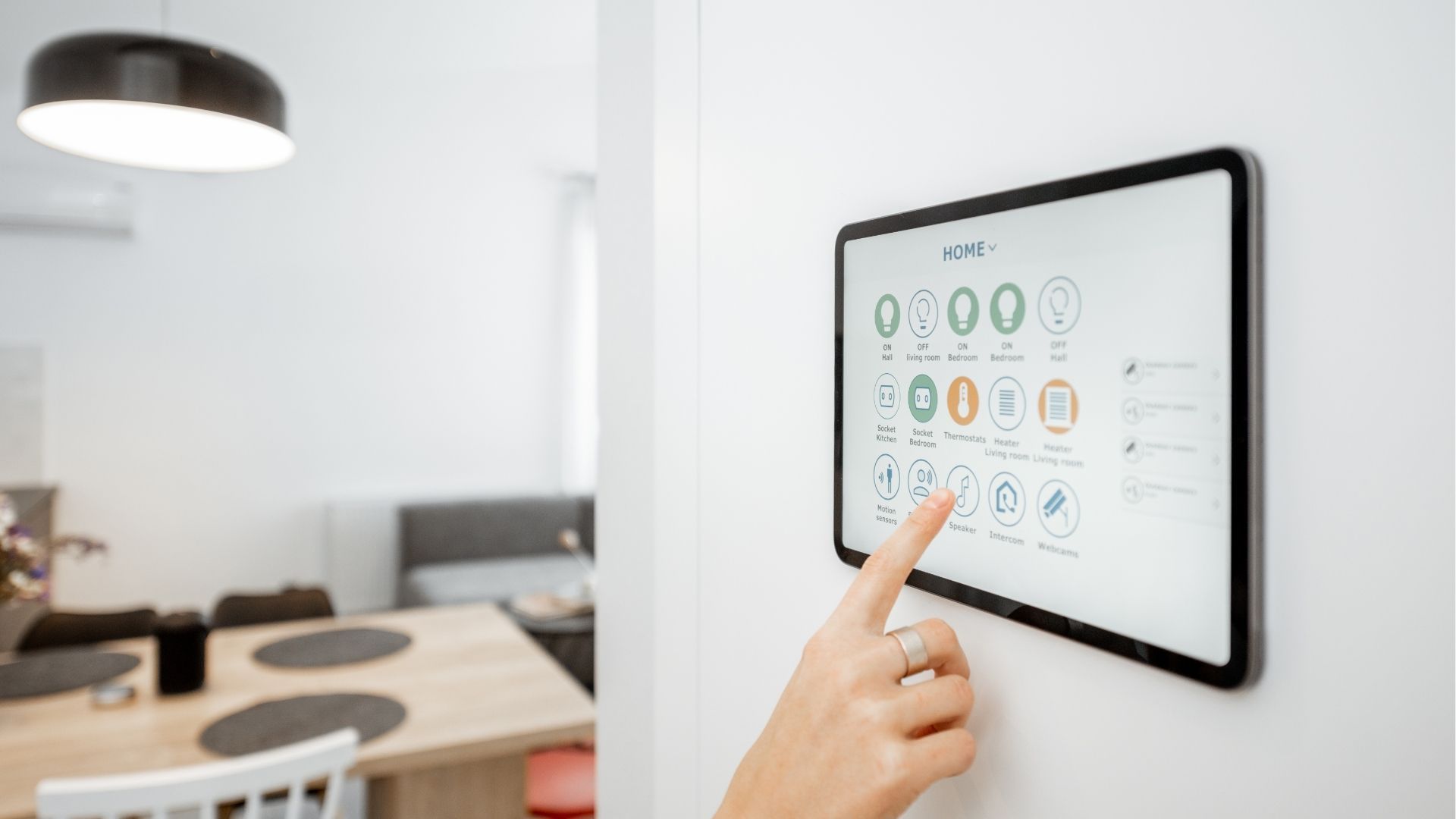 A hand touches a wall-mounted smart home control screen in a modern dining area.