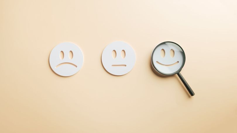 Three circular icons with sad, neutral, and smiling faces on a beige background, with a magnifying glass over the smile.