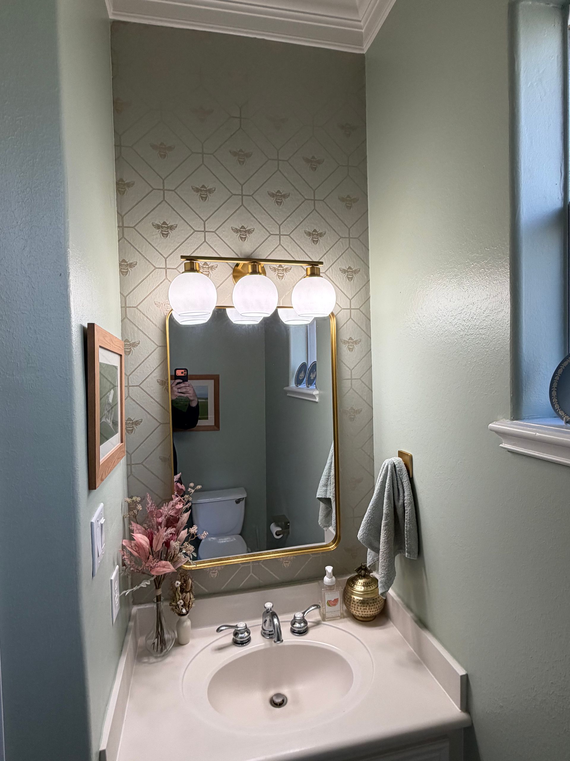 A bathroom vanity with a patterned accent wall, gold-framed mirror, three-globe light fixture, and pale sage green walls.