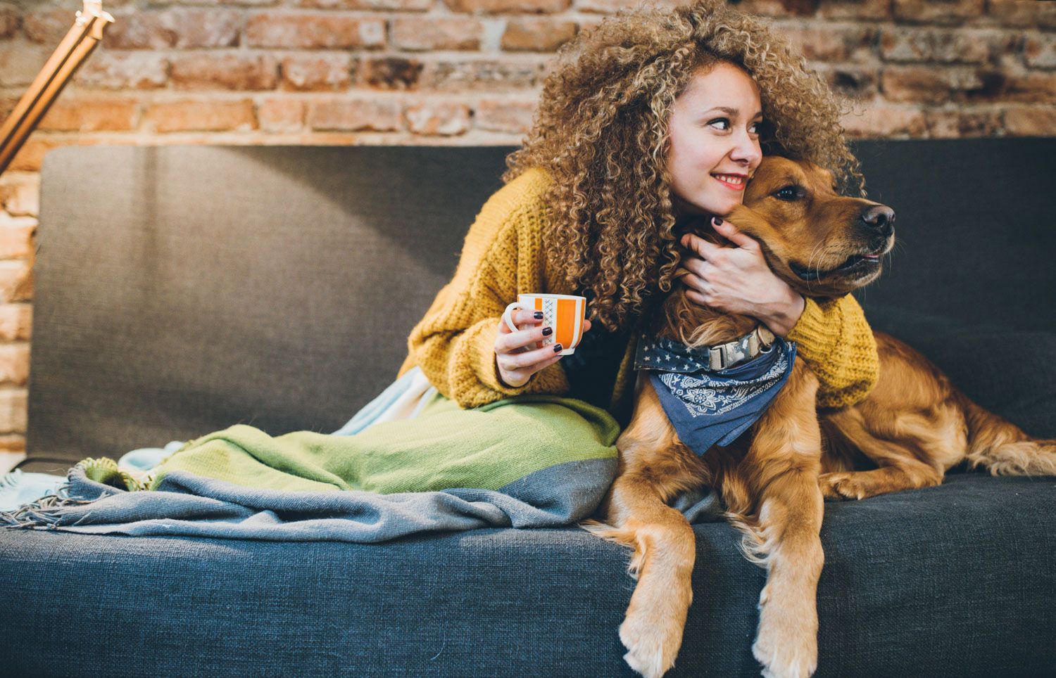Woman on couch hugs golden retriever, holding a mug. Cozy scene, warm tones, and bandana.