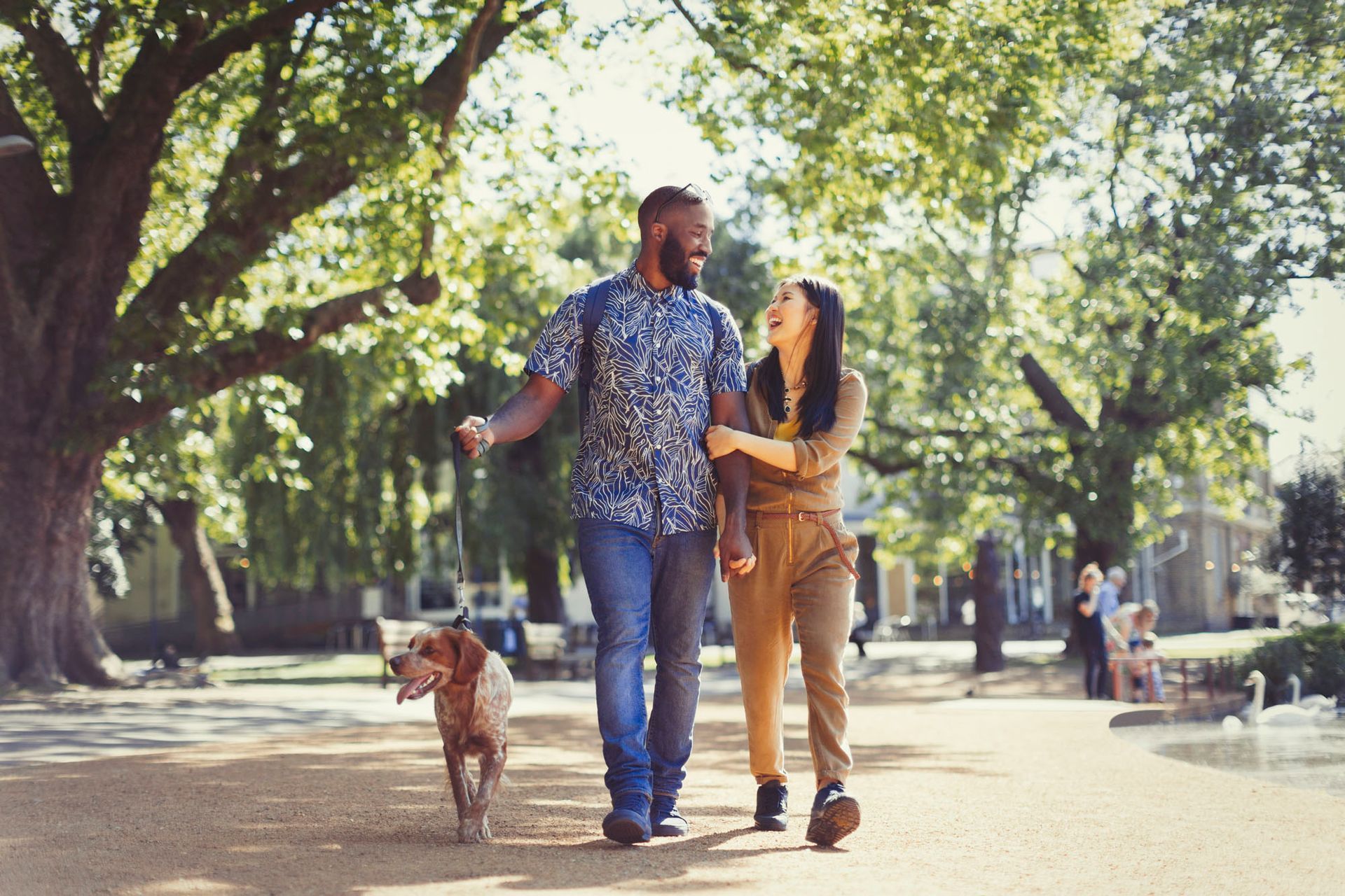 Couple walks dog in park, trees overhead, sunny day. Man laughs, woman smiles.