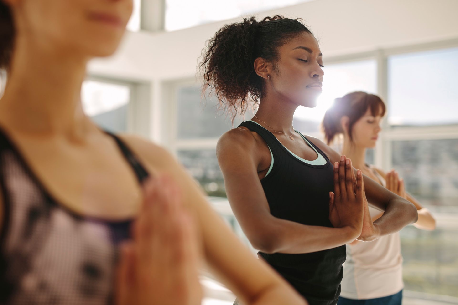 Women in yoga class, hands in prayer pose, eyes closed, bright room setting.