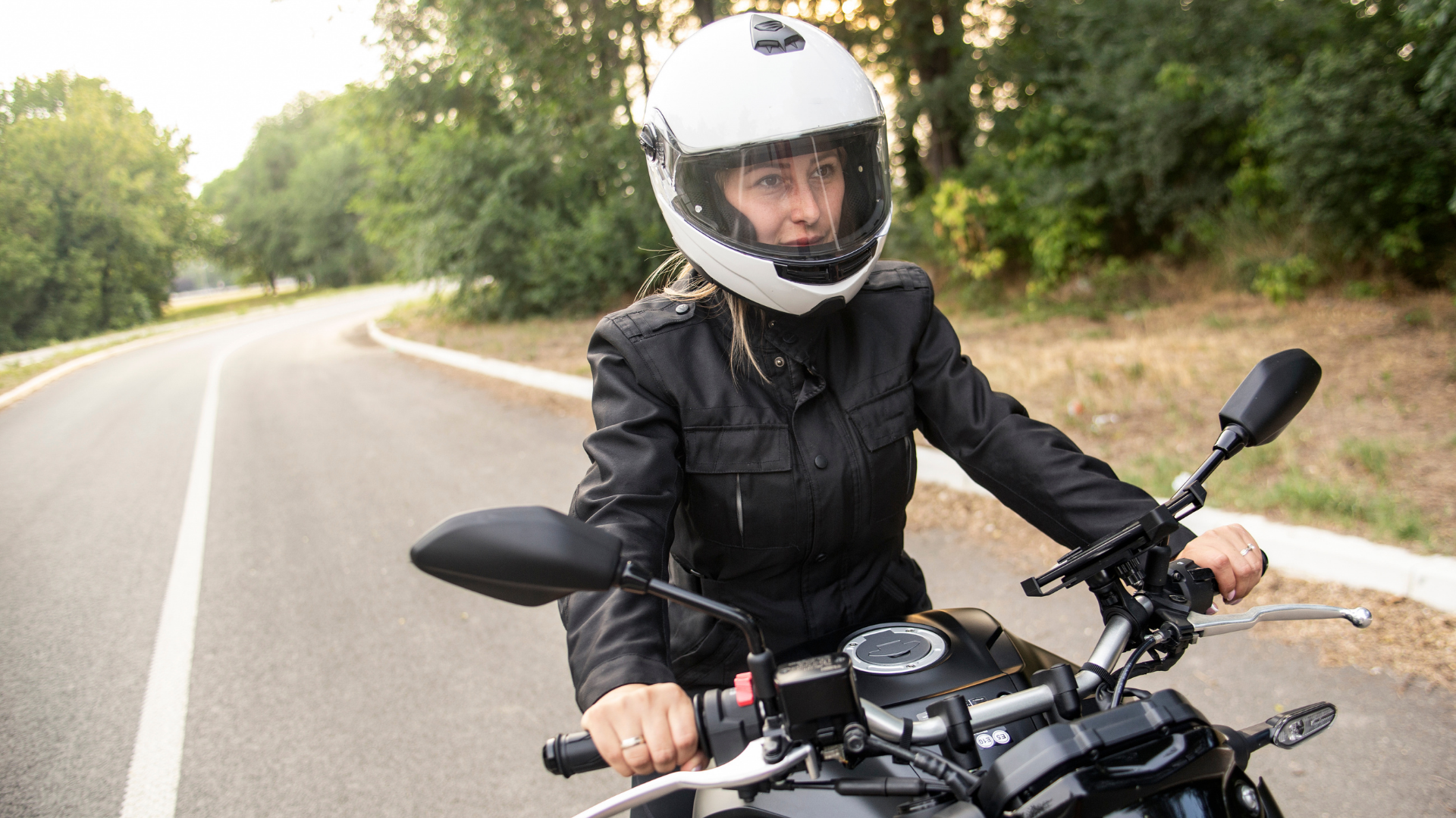 Motorcyclist in black jacket and white helmet riding on a winding road lined with trees.
