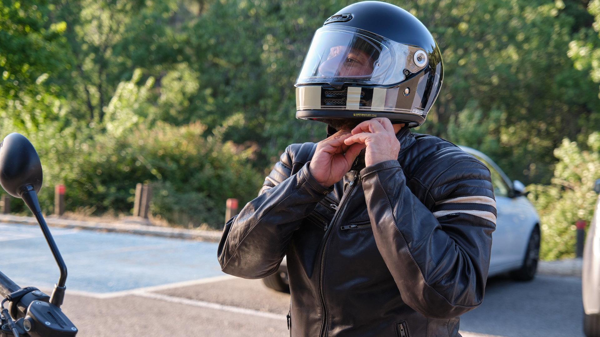 Person in a black motorcycle jacket fastening a helmet outdoors beside a parked car.