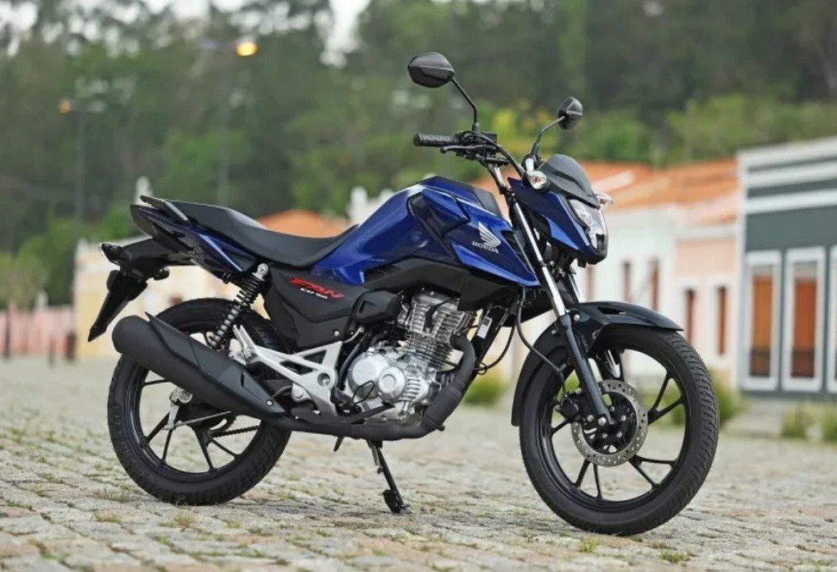 Blue motorcycle parked on a cobblestone path near a building, with trees in the background.