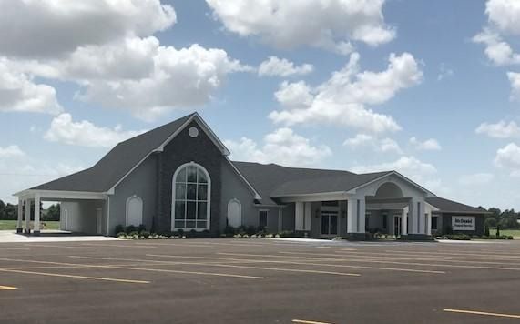 A wide view of a grey commercial building with a steep roof, large arched window, and portico entrance under a cloudy sky.