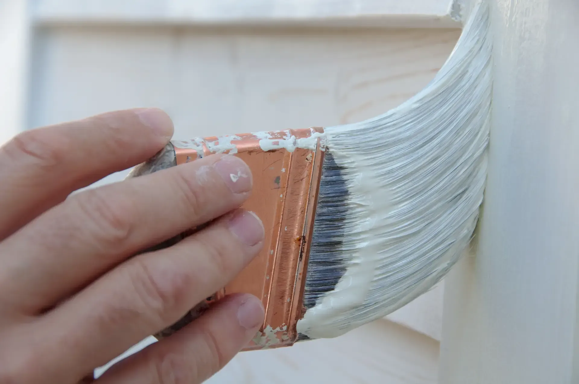 Hand holding a paint brush, applying white paint to a wooden surface.