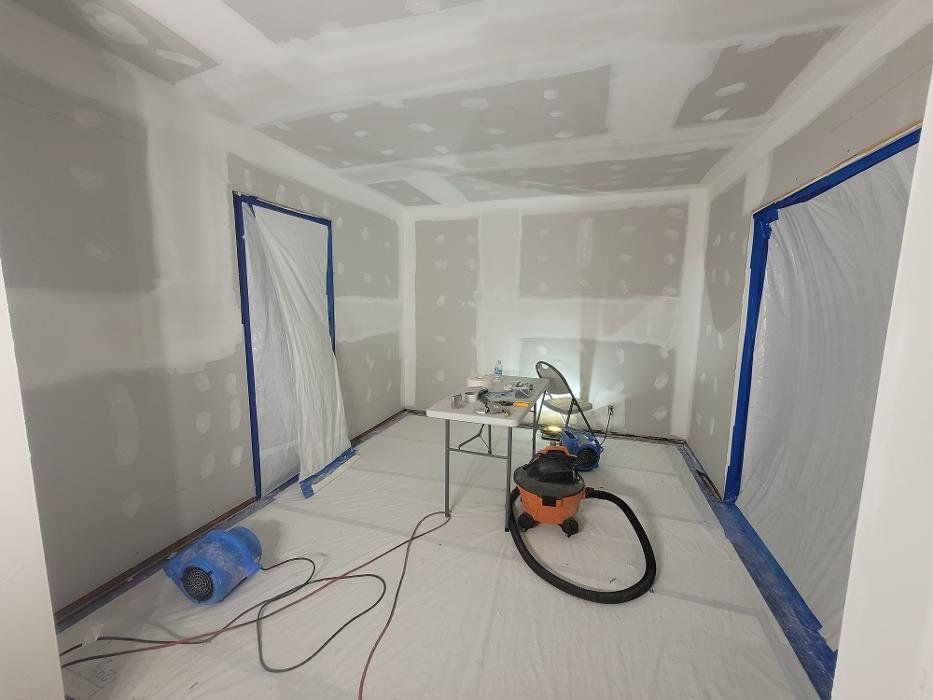 Room under renovation; drywall, tape, and plastic sheeting protect surfaces. Table with tools, vacuum cleaner.