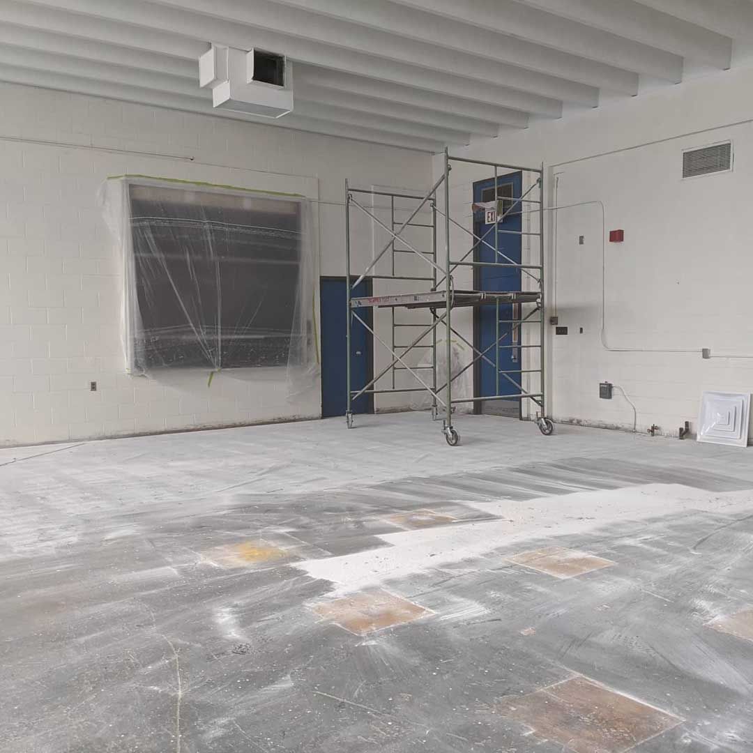 Interior construction site with scaffolding, unfinished floor, boarded-up window, and exposed ceiling.