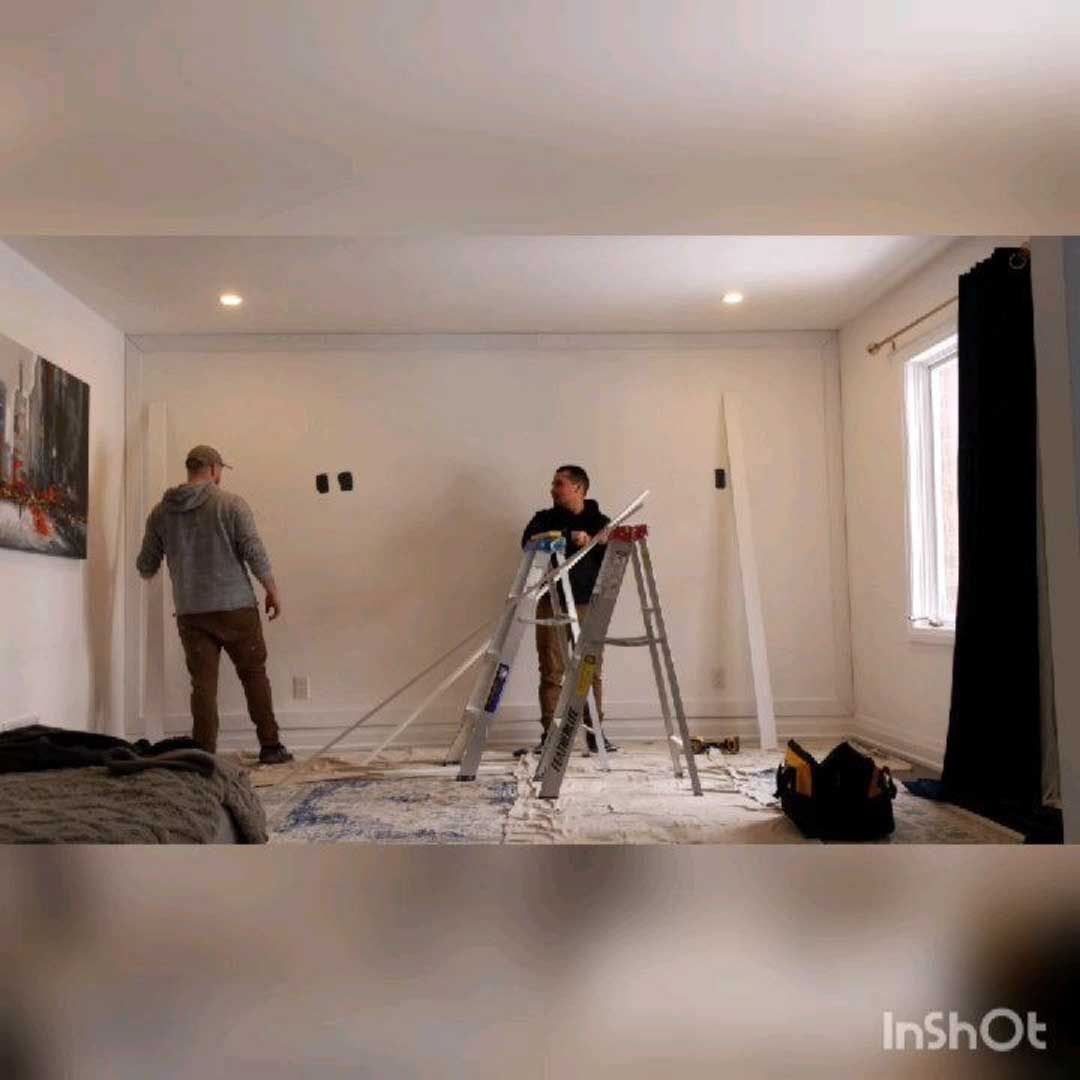 Two people renovating a room; one on a ladder, the other standing. White walls, tools, and debris are visible.