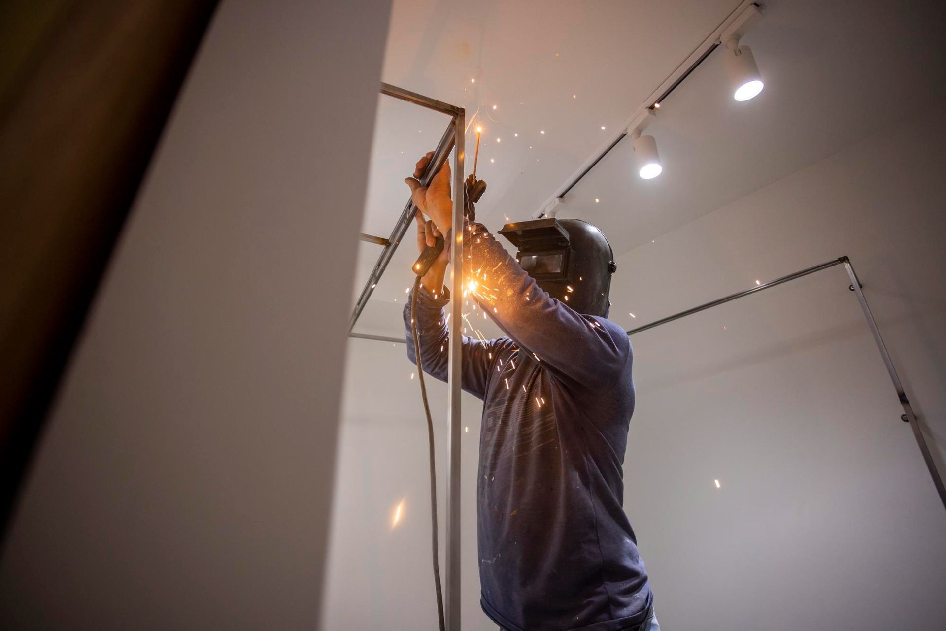 Welder wearing a helmet working on a metal frame, creating sparks in an indoor setting.