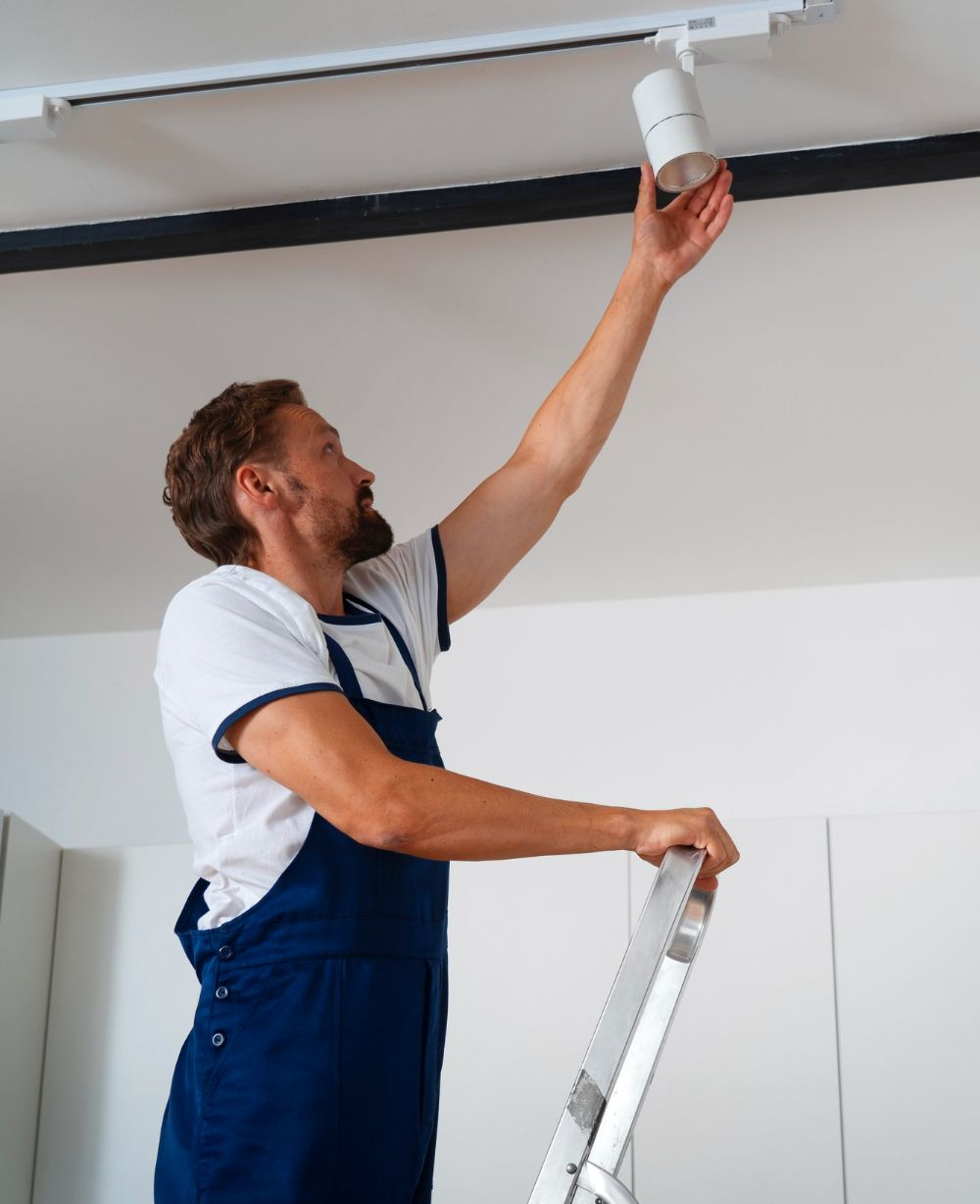 Man on a ladder installing a cylindrical light fixture on a track in a white ceiling.