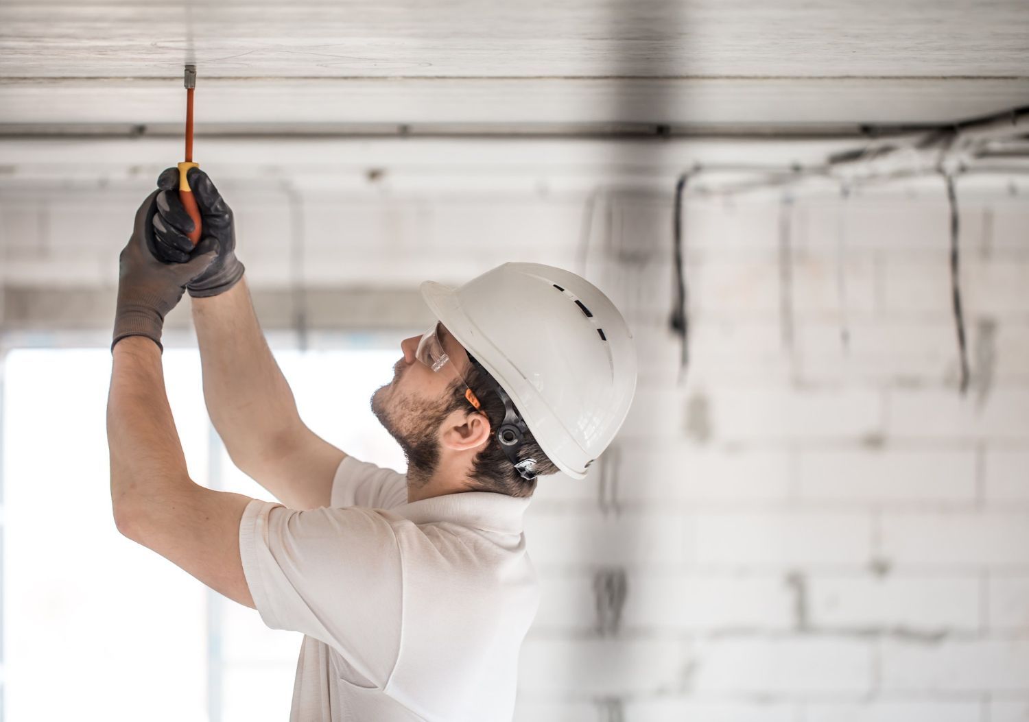 Construction worker in white hard hat and gloves working on ceiling with a screwdriver.