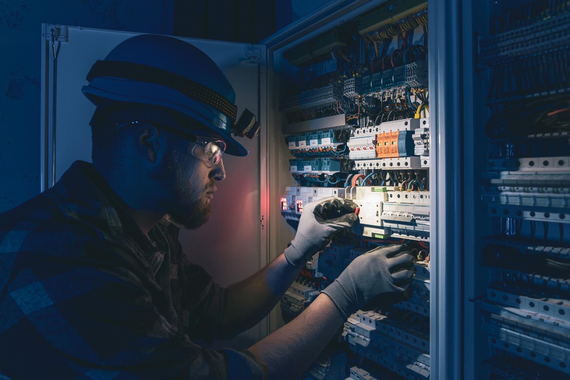Electrician working on electrical panel, wearing a hard hat with a headlamp and safety gloves.