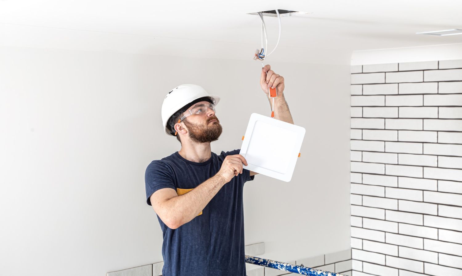 Electrician in a hard hat working on ceiling wiring, holding a plan in a room with white brick wall.