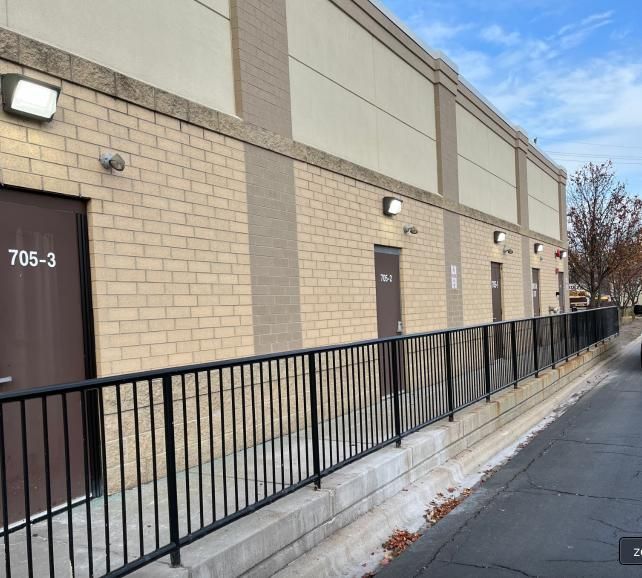 Exterior of a brick building with dark doors labeled with numbers. Black railing runs along a concrete base.