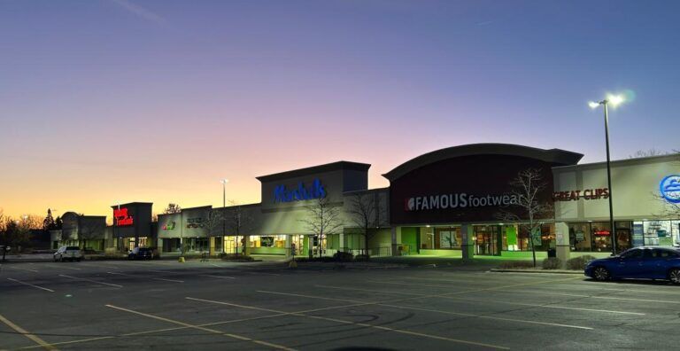 Shopping plaza exterior at dusk with several lit store signs against a purple and orange sky.
