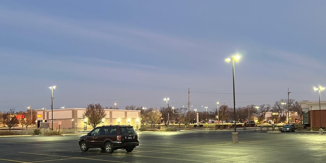 Dark SUV driving through empty parking lot at dusk, with streetlights illuminating the scene.