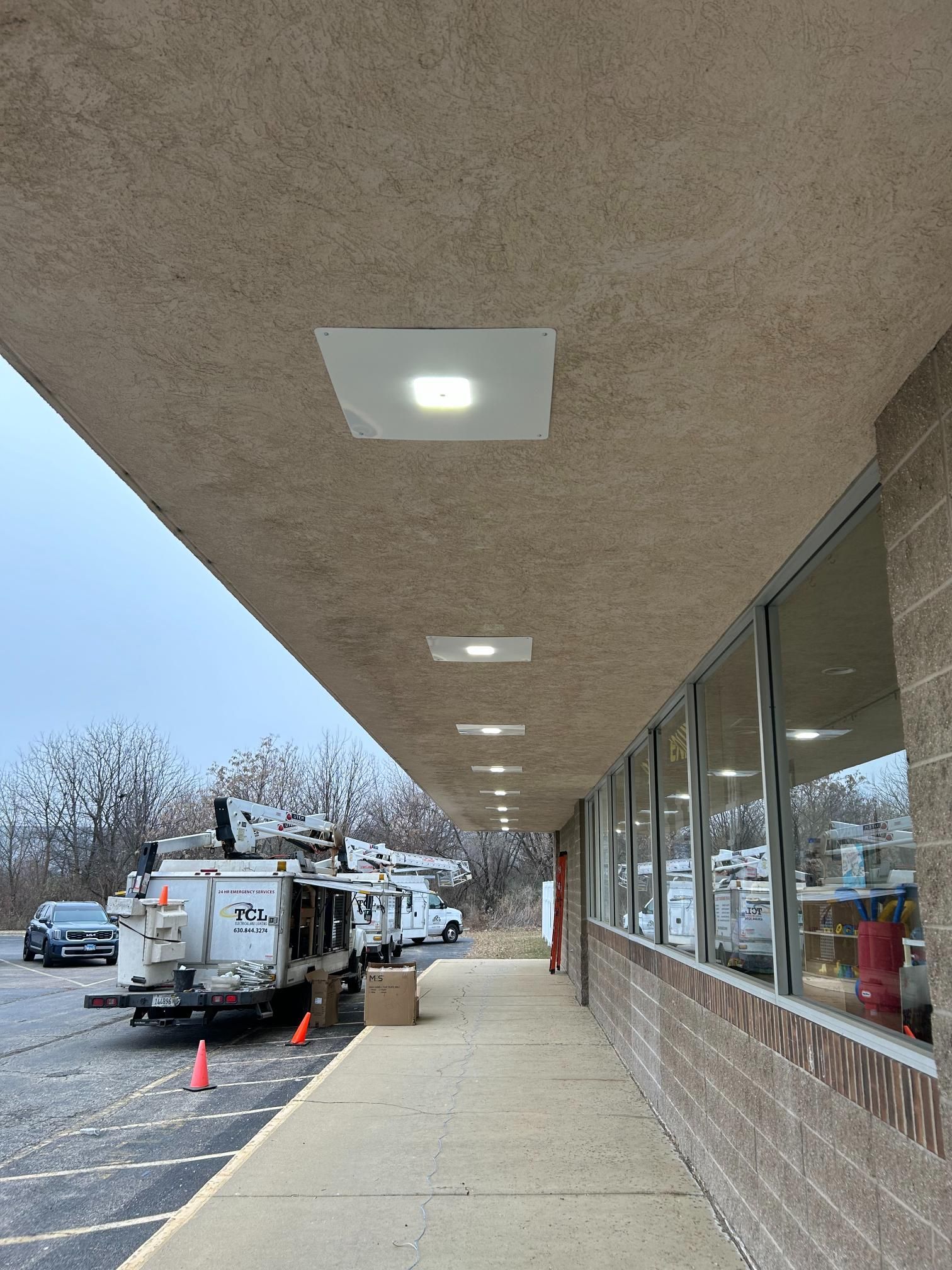 Exterior of a building with overhead lighting and a row of windows. Construction vehicles are parked nearby.