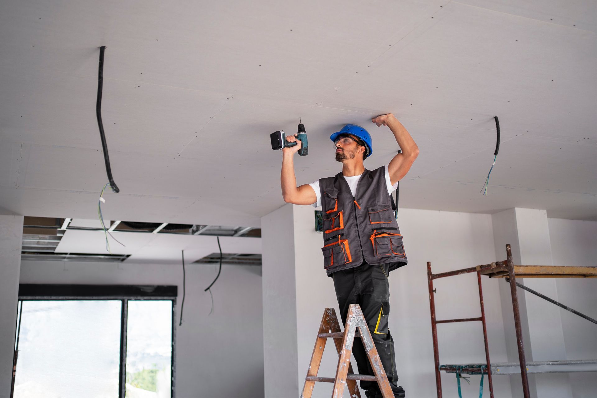 Construction worker on a ladder drills into the ceiling. Wires hang; interior space.