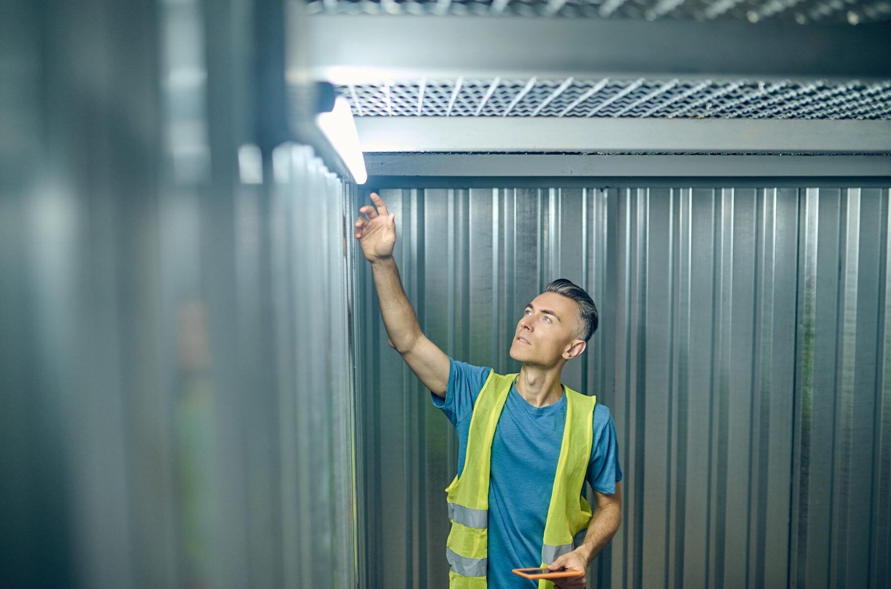 Man in a yellow vest examines a light fixture in a metal-walled storage unit.