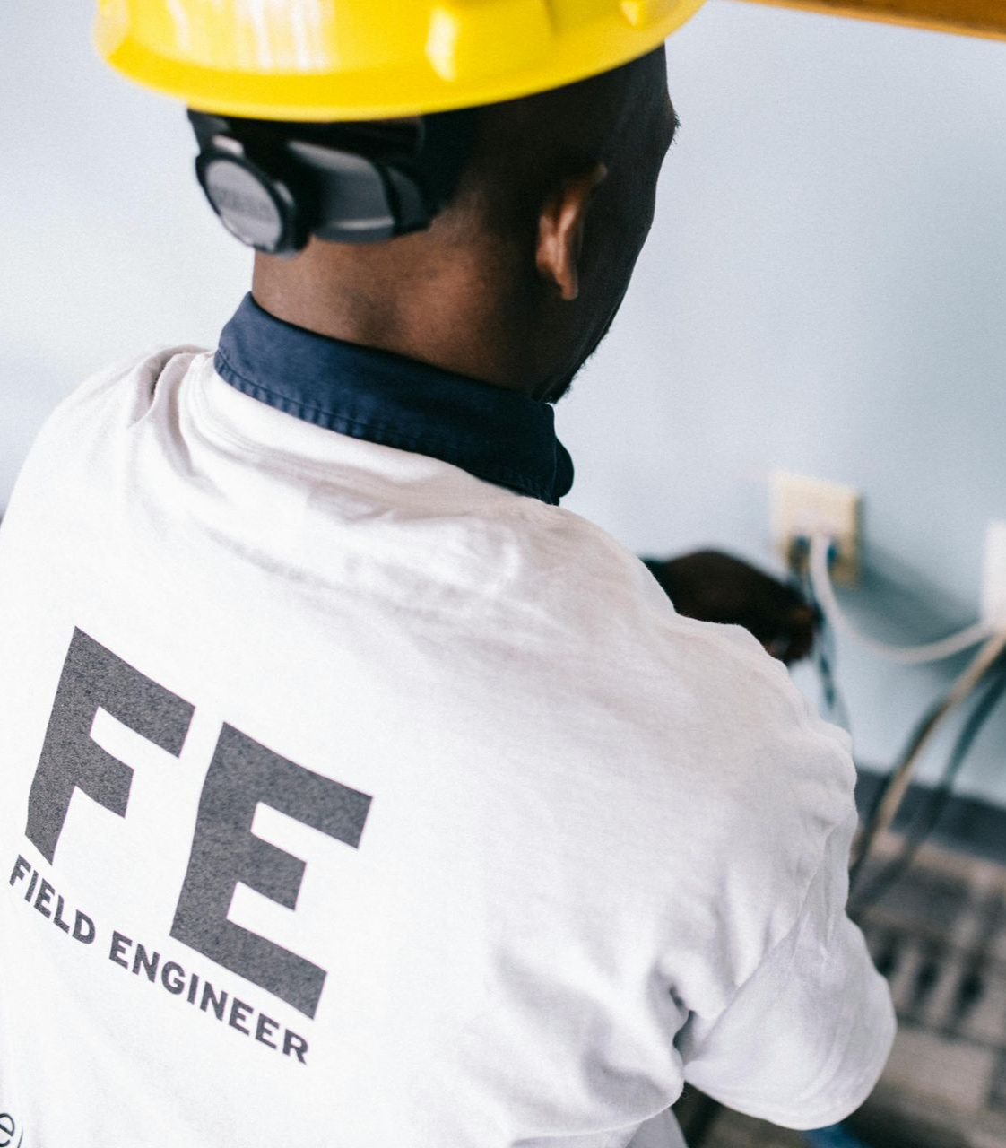 Field engineer in yellow hard hat working with wires at wall outlets. White shirt with