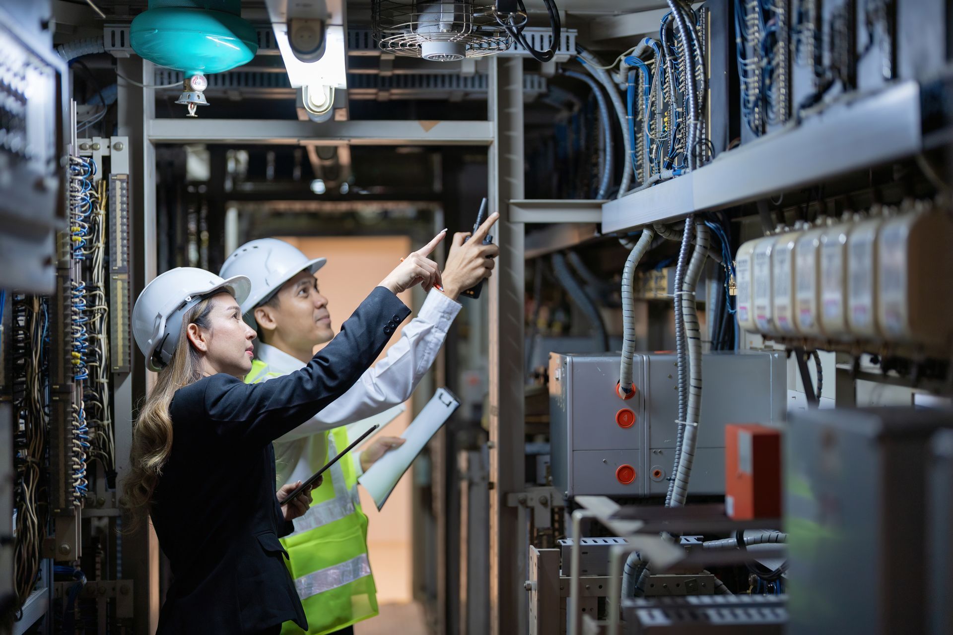 Two people in hard hats inspect electrical equipment, pointing.